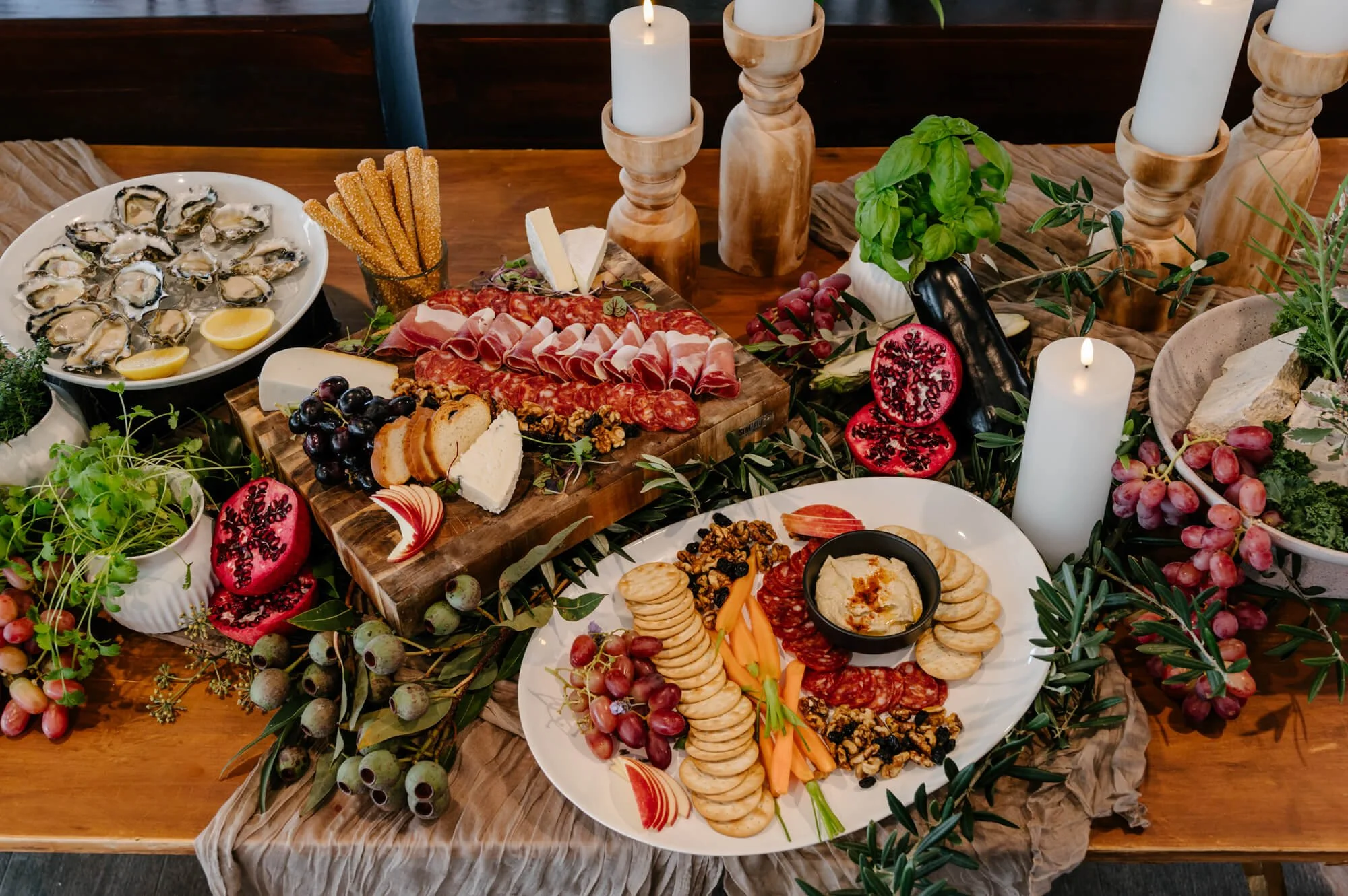 A wooden table set with various cheeses, cured meats, fresh fruit, crackers, oysters, and candles for a festive gathering.