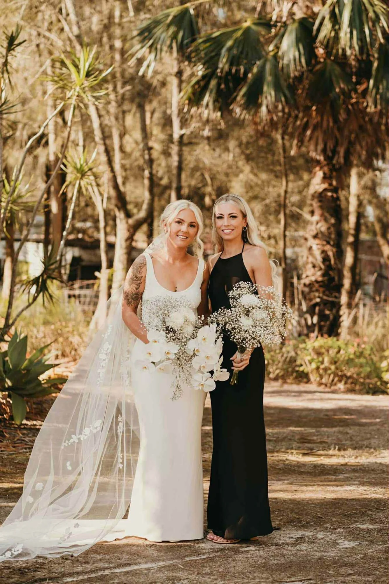 Two women, a bride in a white wedding dress with a veil and floral bouquet, and a bridesmaid in a black dress with a bouquet, standing outdoors among trees and greenery.