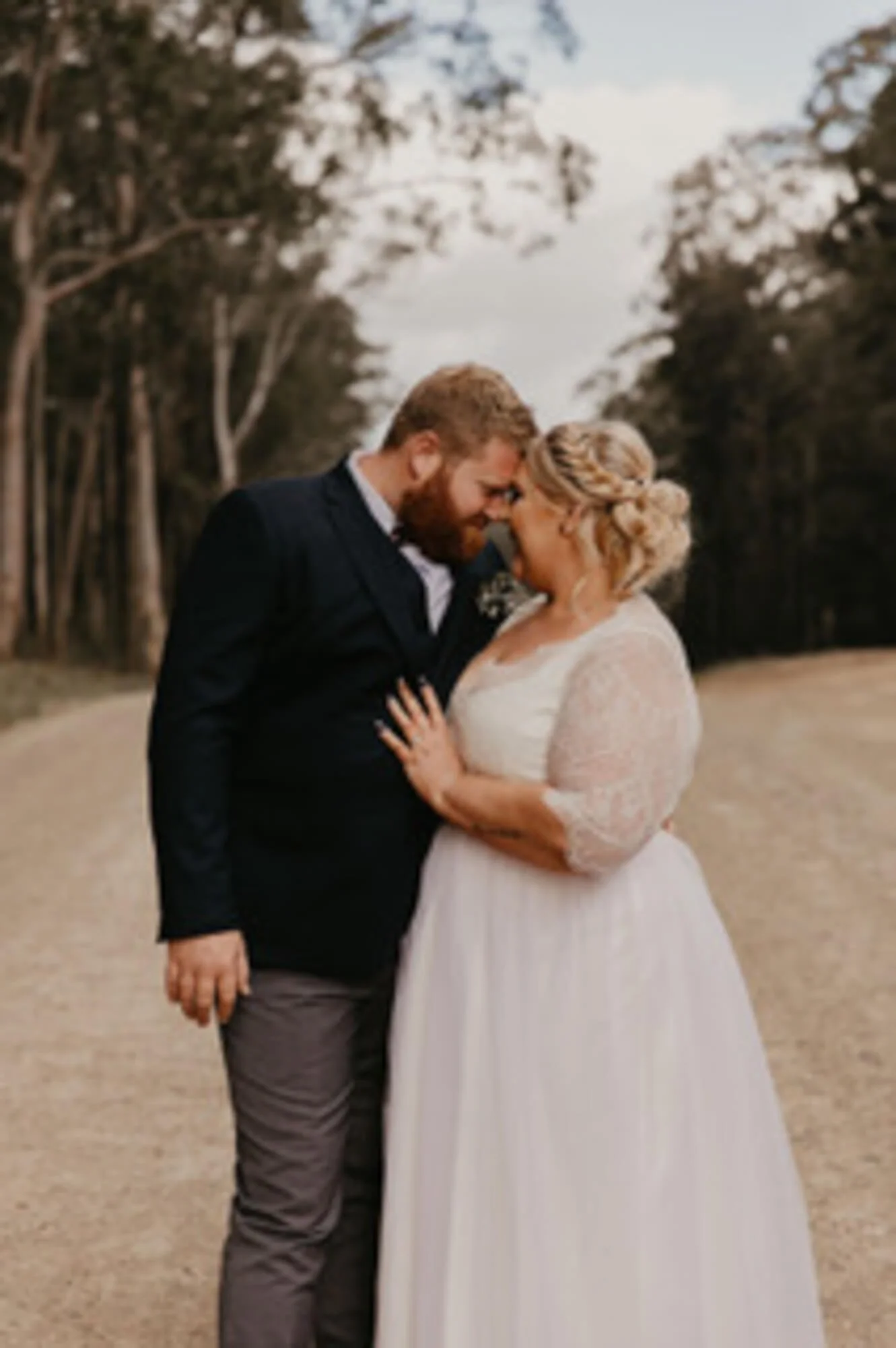 A couple on their wedding day, standing closely with foreheads touching, outdoors with trees and a cloudy sky in the background.