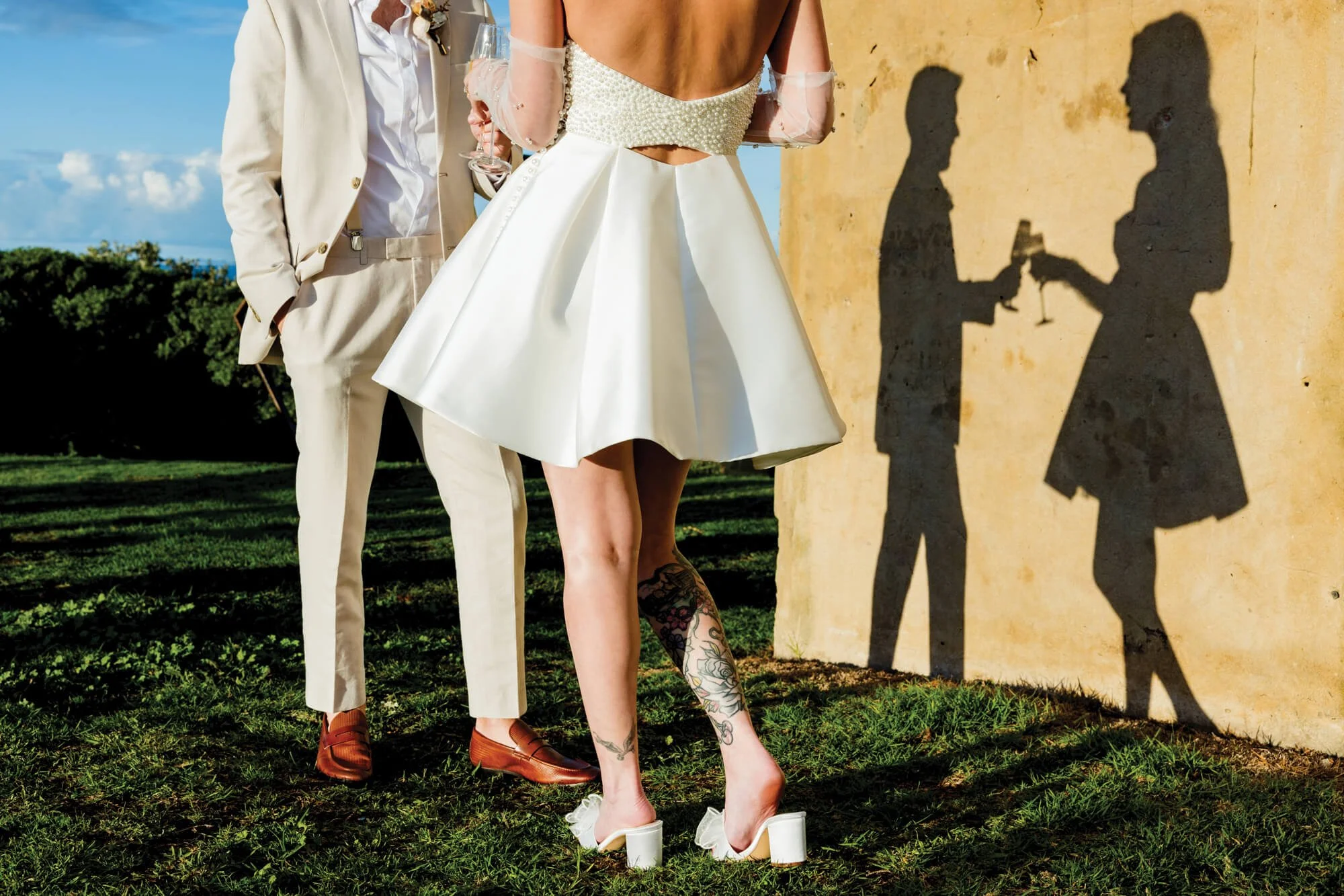 Shadow of a couple holding champagne glasses and a toast on a wall, with a woman wearing a white dress and tattooed leg and man in a light beige suit standing on grass background.