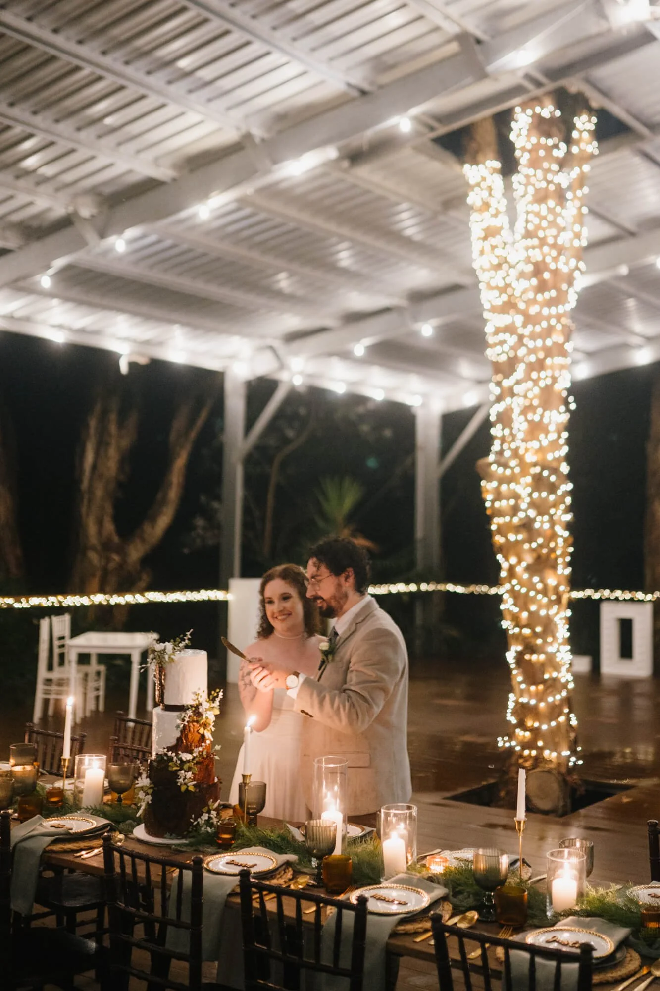 A couple celebrating a wedding or special occasion in an outdoor setting at night, with string lights and a tree wrapped in fairy lights in the background. They are standing near a decorated table with candles and a cake.