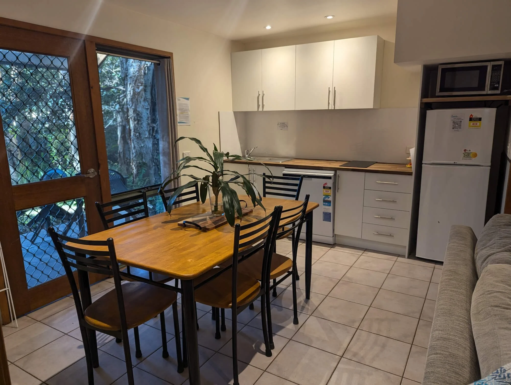 Kitchen with dining table, black chairs, white cabinets, refrigerator, microwave, and large window showing outdoor greenery.