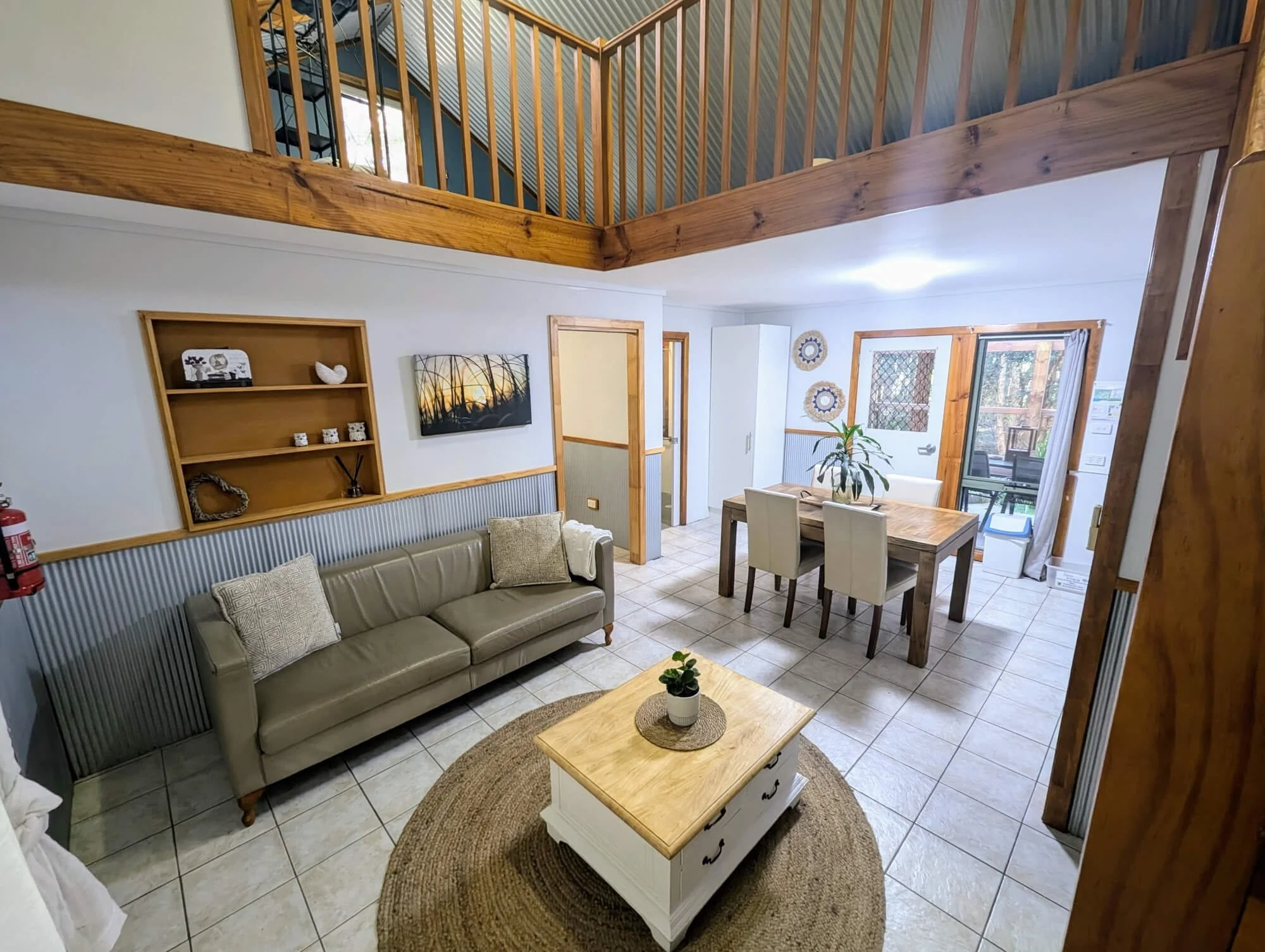 Interior of a living and dining area with a leather sofa, wooden coffee table, dining table with chairs, a railing on the second level, and large windows with outdoor views.