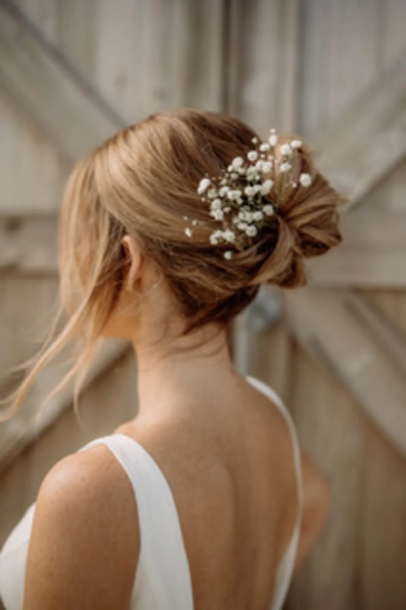 Back of a woman with styled hair adorned with white baby's breath flowers, wearing a white dress, against a wooden background.