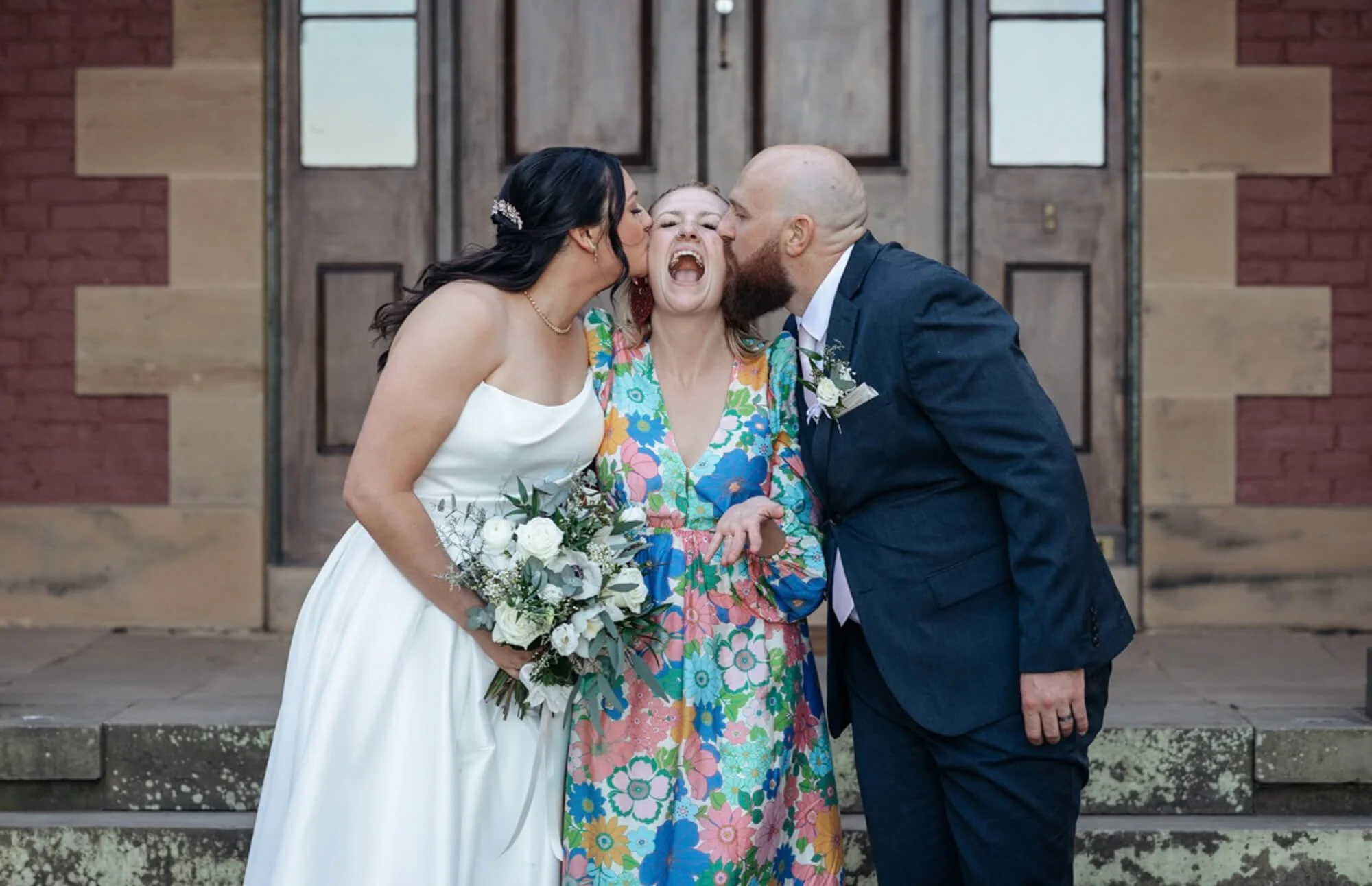 A bride in a white wedding dress and a groom in a navy suit kiss a woman in a colorful floral dress on each cheek during a wedding celebration outside a building with wooden doors.