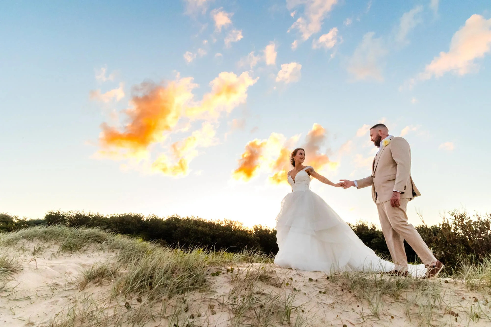 A bride and groom holding hands on a sandy beach during sunset, with orange and pink clouds in the sky.