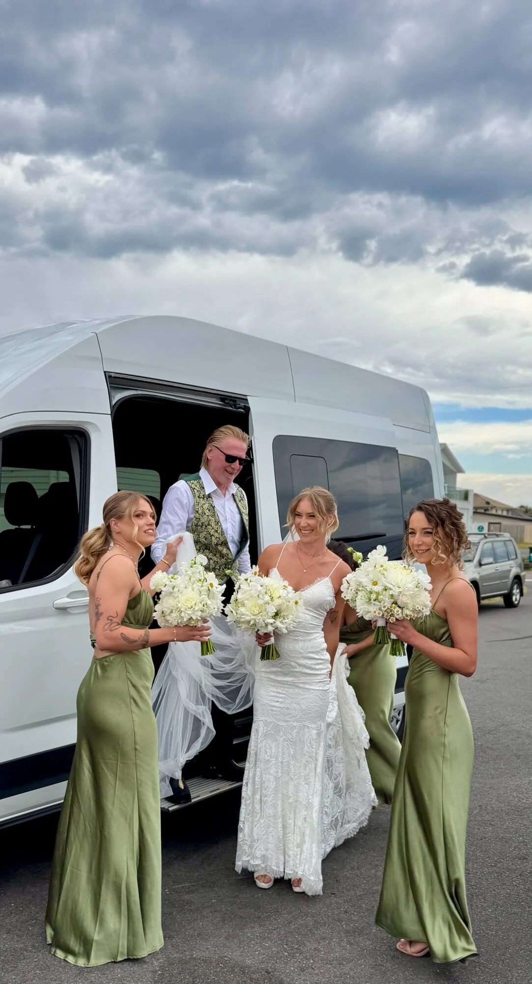 A bride in a white wedding dress with lace details and three bridesmaids in matching green satin dresses standing outside near a white limousine with a group of people. The sky is cloudy, and it appears to be late afternoon or early evening.