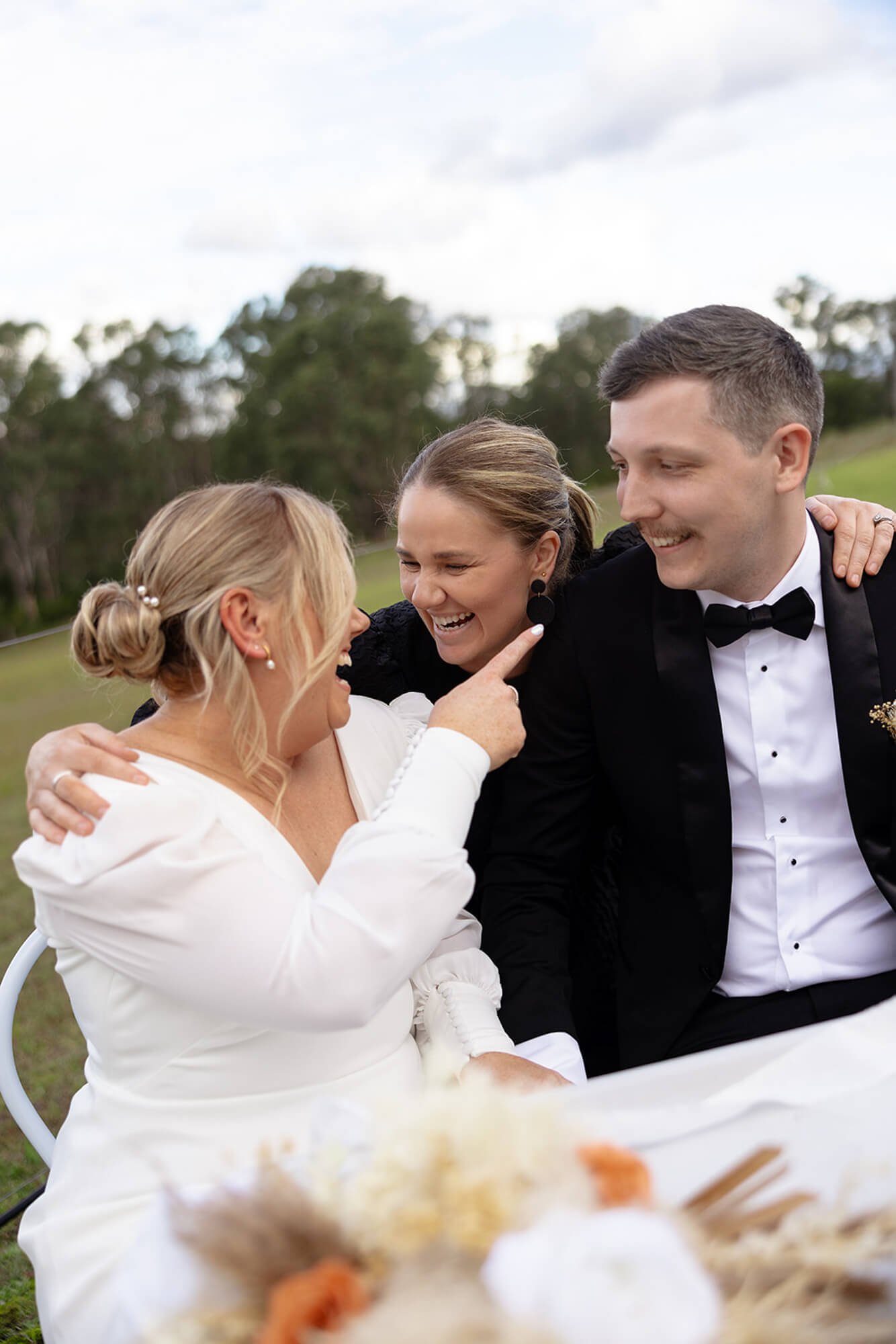 Three people at a wedding, an older woman with blonde hair, a woman with light brown hair, and a man with short dark hair, all smiling and laughing outdoors with trees in the background.