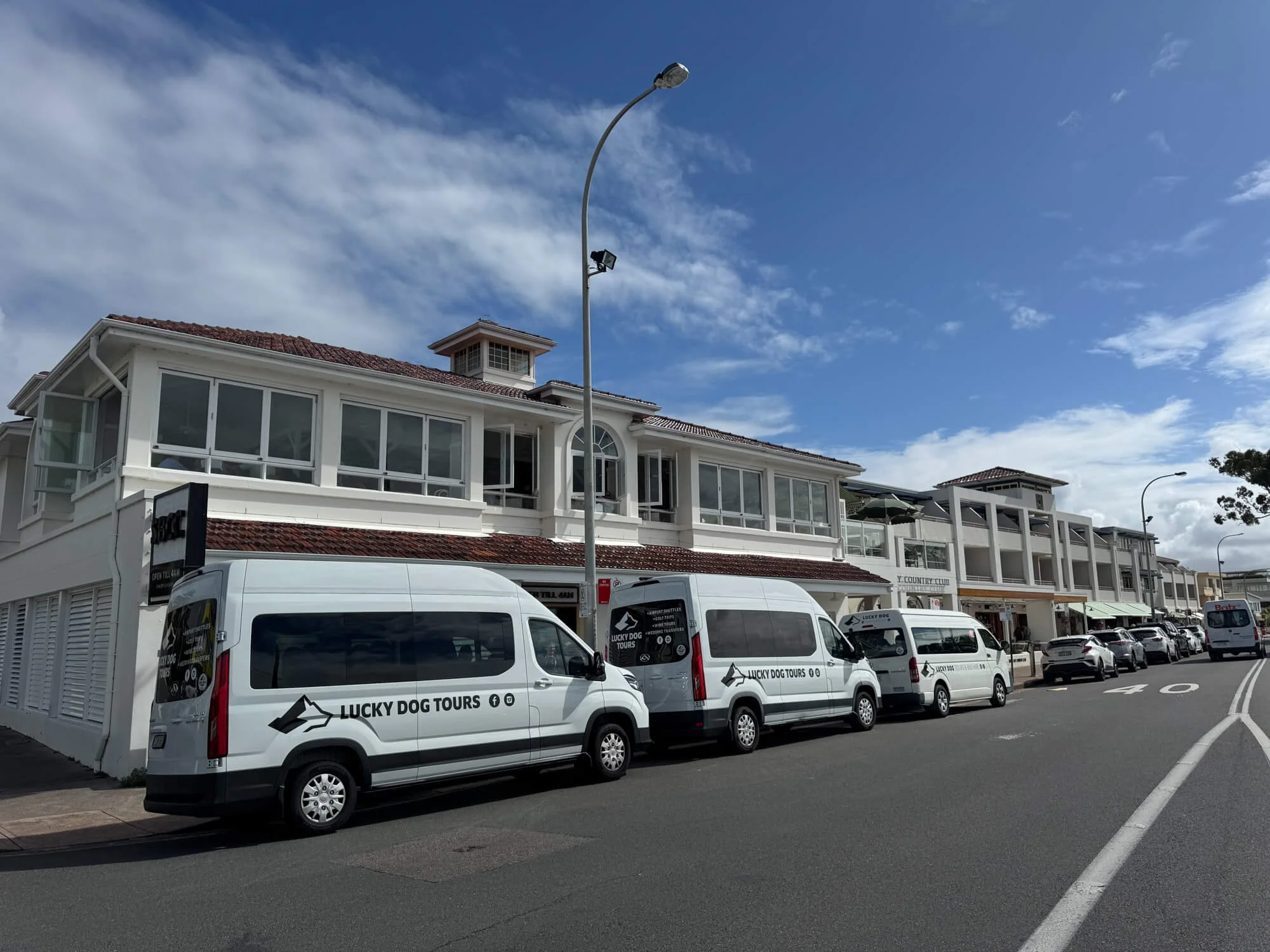 Street view with parked vans and cars in front of a two-story building with large windows, under partly cloudy sky.
