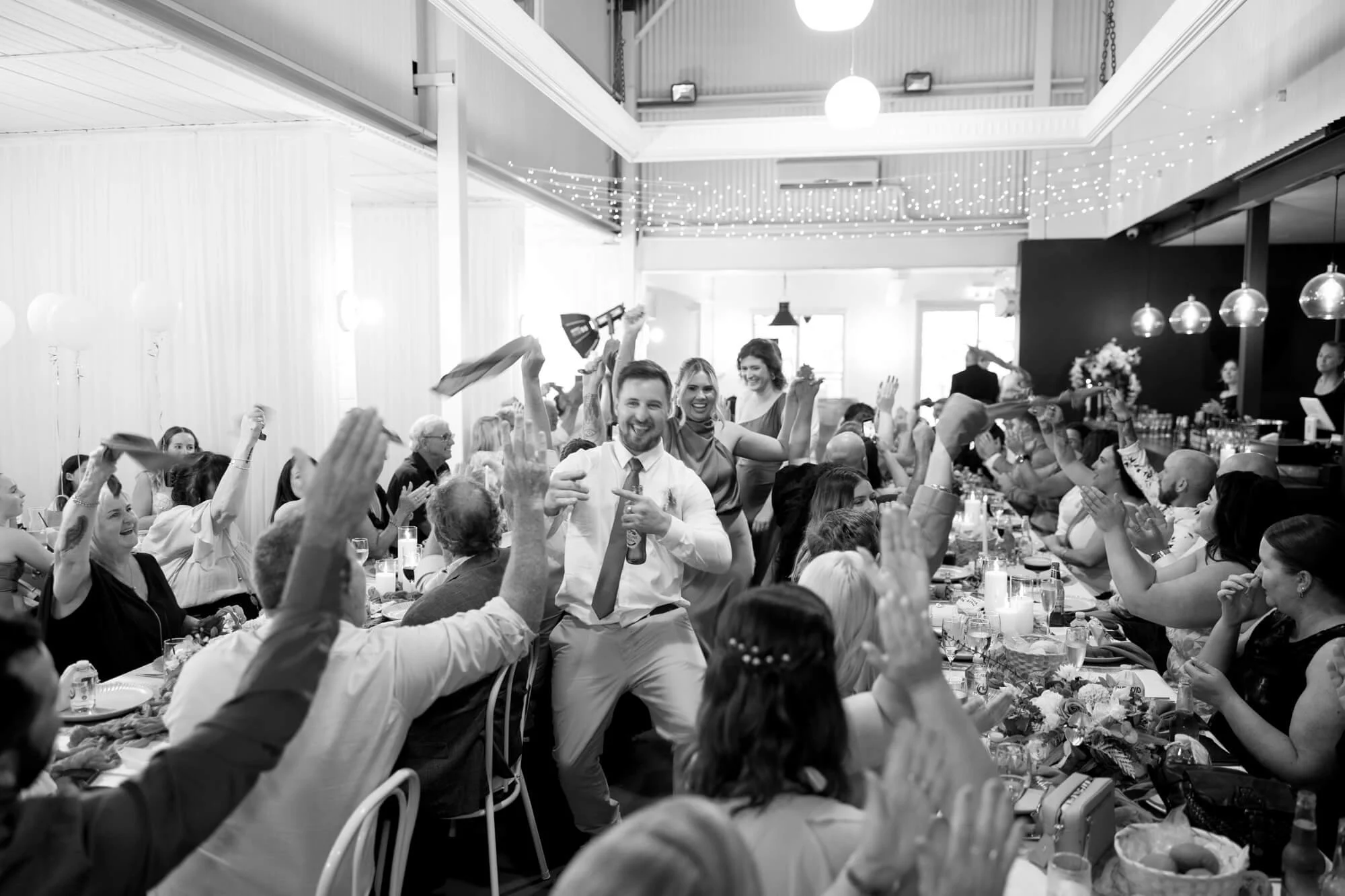 People celebrating at a wedding reception, with one man dancing and pointing, surrounded by guests clapping and cheering at a long decorated table.