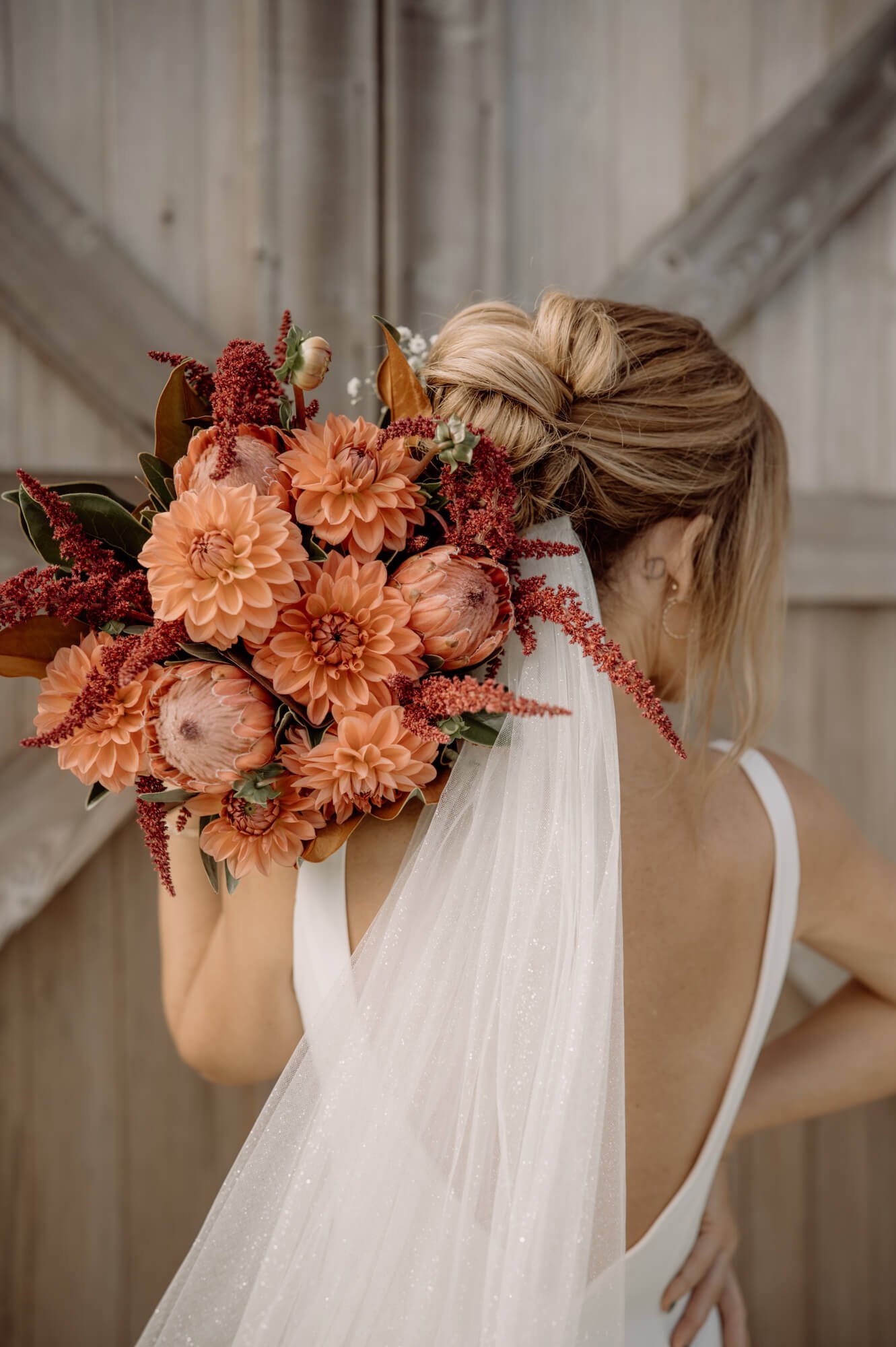 A bride holding a large peach and red flower bouquet, wearing a white dress and a sheer veil, with her hair in a loose bun. The background features a wooden wall with diagonal beams.