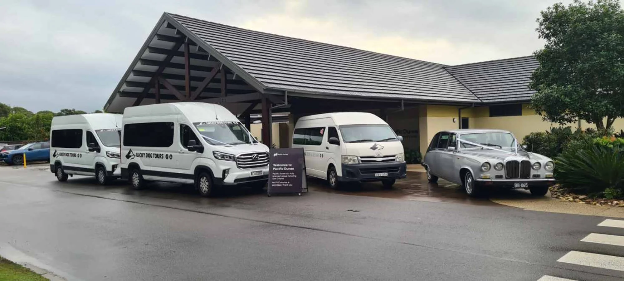 Three tour vans parked outside a building, with a classic silver vintage car parked on the right side, on a rainy day.