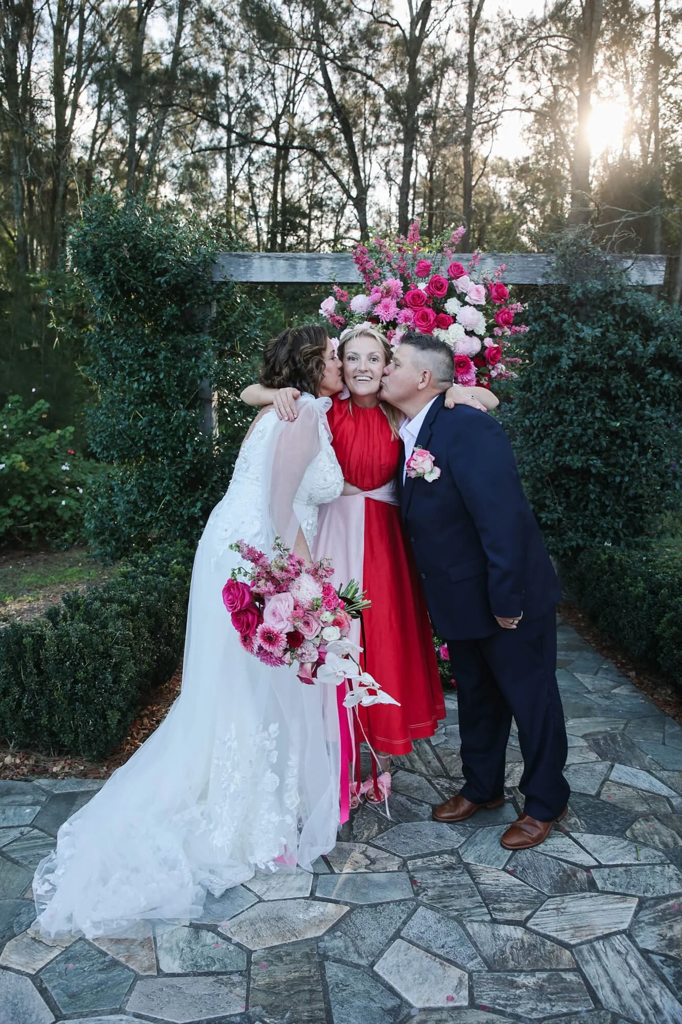 A wedding scene with two women and a man outdoors on a stone pathway, with green bushes and trees, and a large floral arrangement of pink and white flowers. The woman on the left wears a white wedding gown and holds a bouquet, the woman in the middle