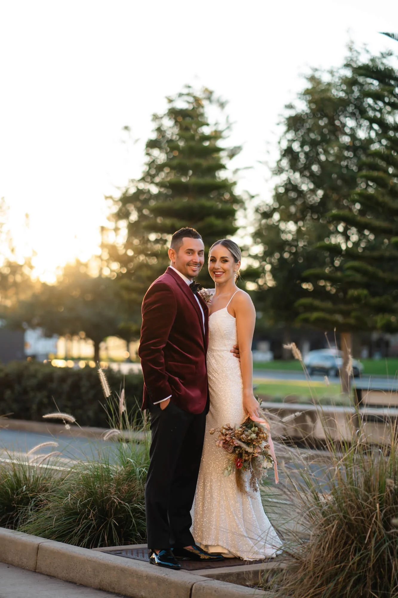 A newlywed couple standing outside at sunset, smiling at the camera. The groom is wearing a burgundy velvet suit jacket and black pants, and the bride is in a white lace wedding dress, holding a bouquet of flowers.