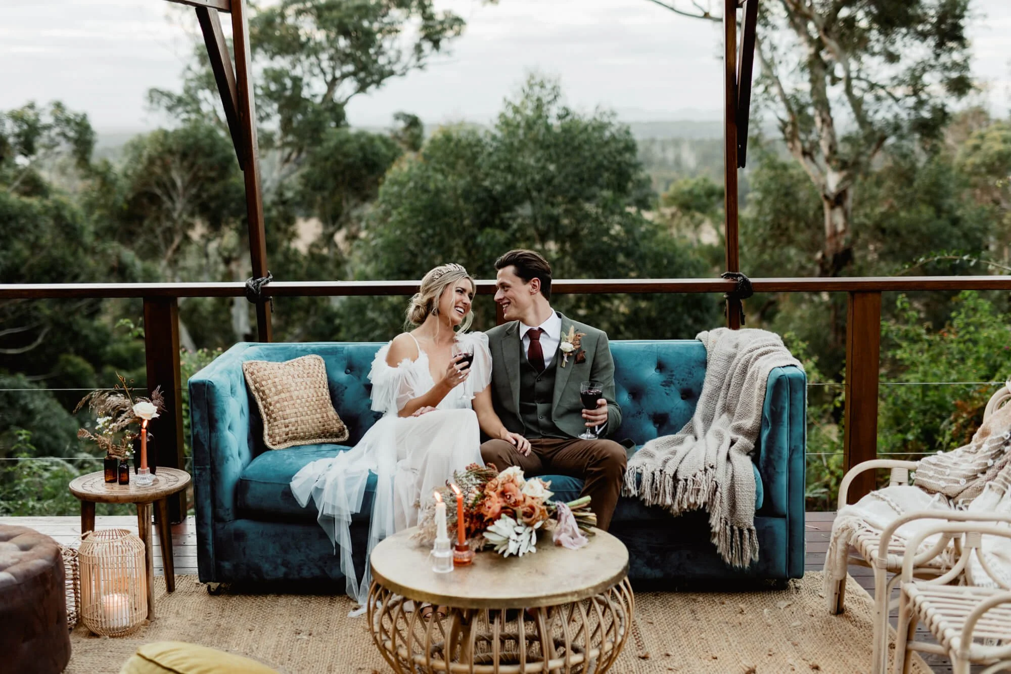 A couple sitting on a blue vintage sofa in an outdoor setting, smiling and looking at each other while holding glasses of red wine. The woman is wearing a white dress with ruffled sleeves and the man is in a dark suit with a boutonniere. There are ca