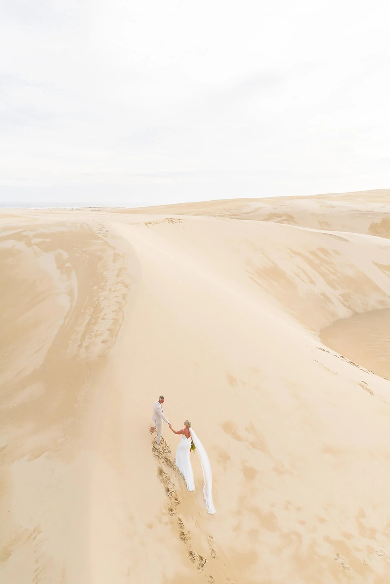 A couple in wedding attire holding hands and walking across a sandy desert with dunes under a cloudy sky.