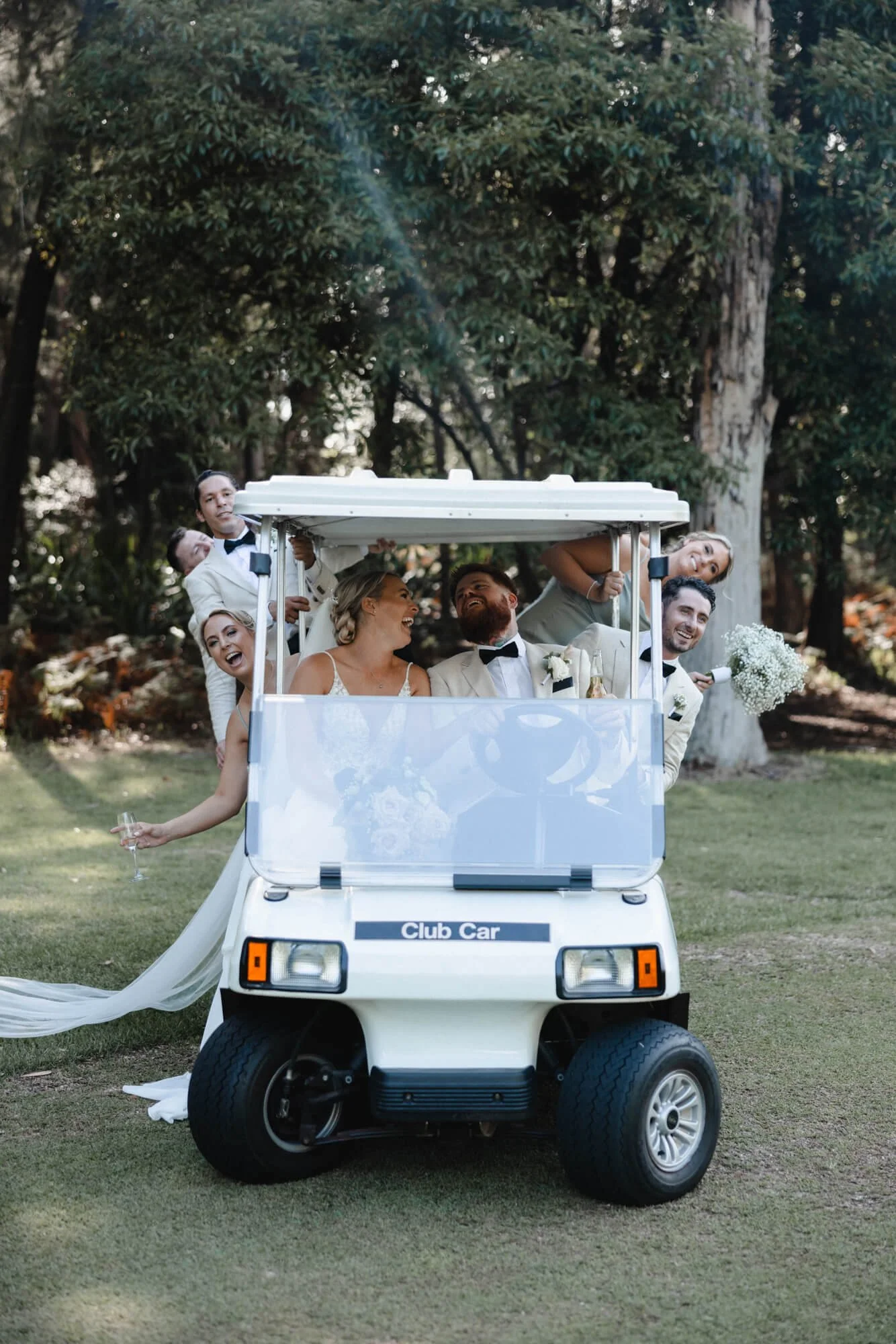 A wedding celebration with a bride, groom, and friends riding in a small white golf cart on a grassy area surrounded by trees.