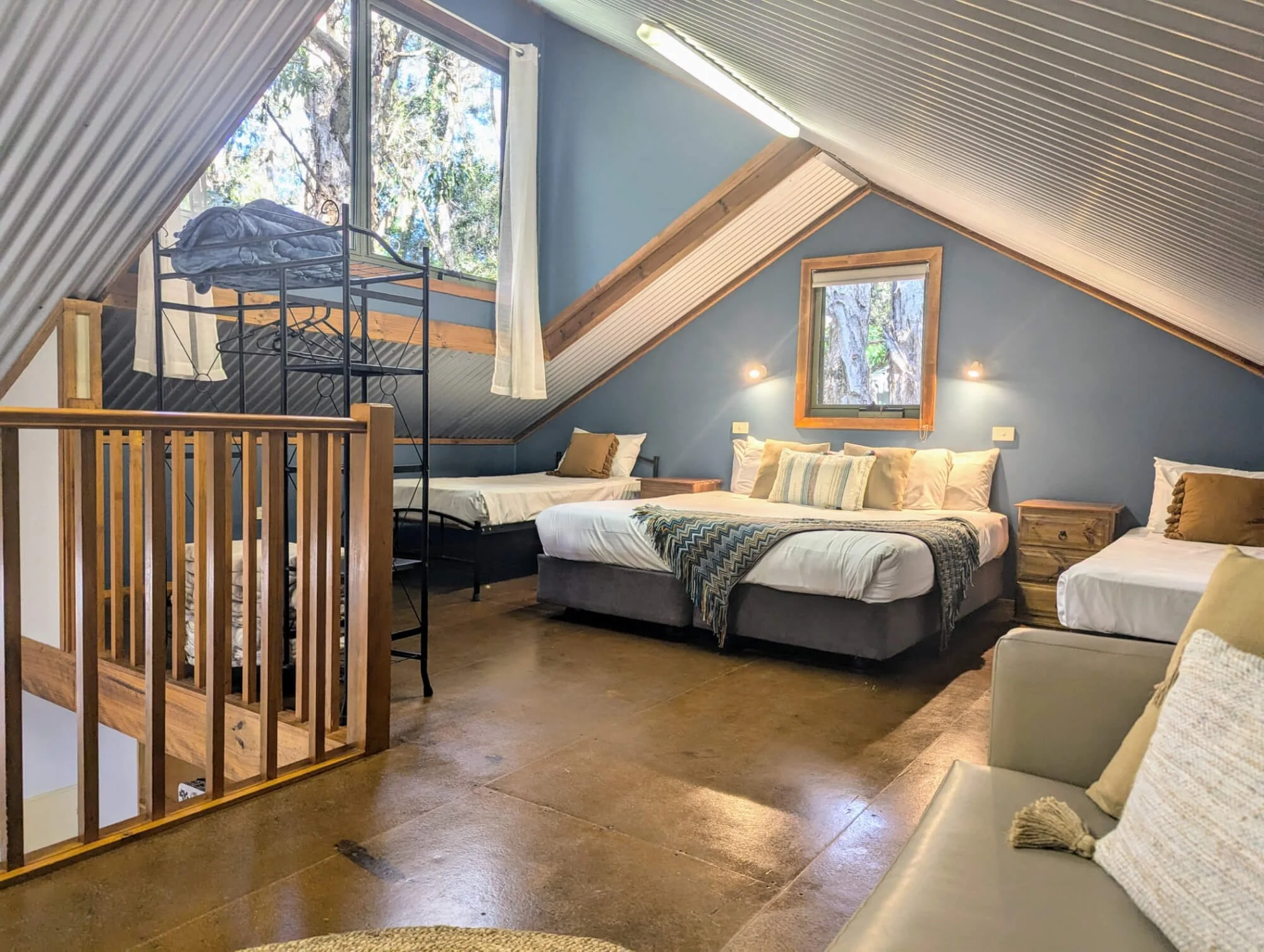 Interior of an attic bedroom with sloped ceiling, featuring three beds, windows showing outdoor trees, and wooden furniture.