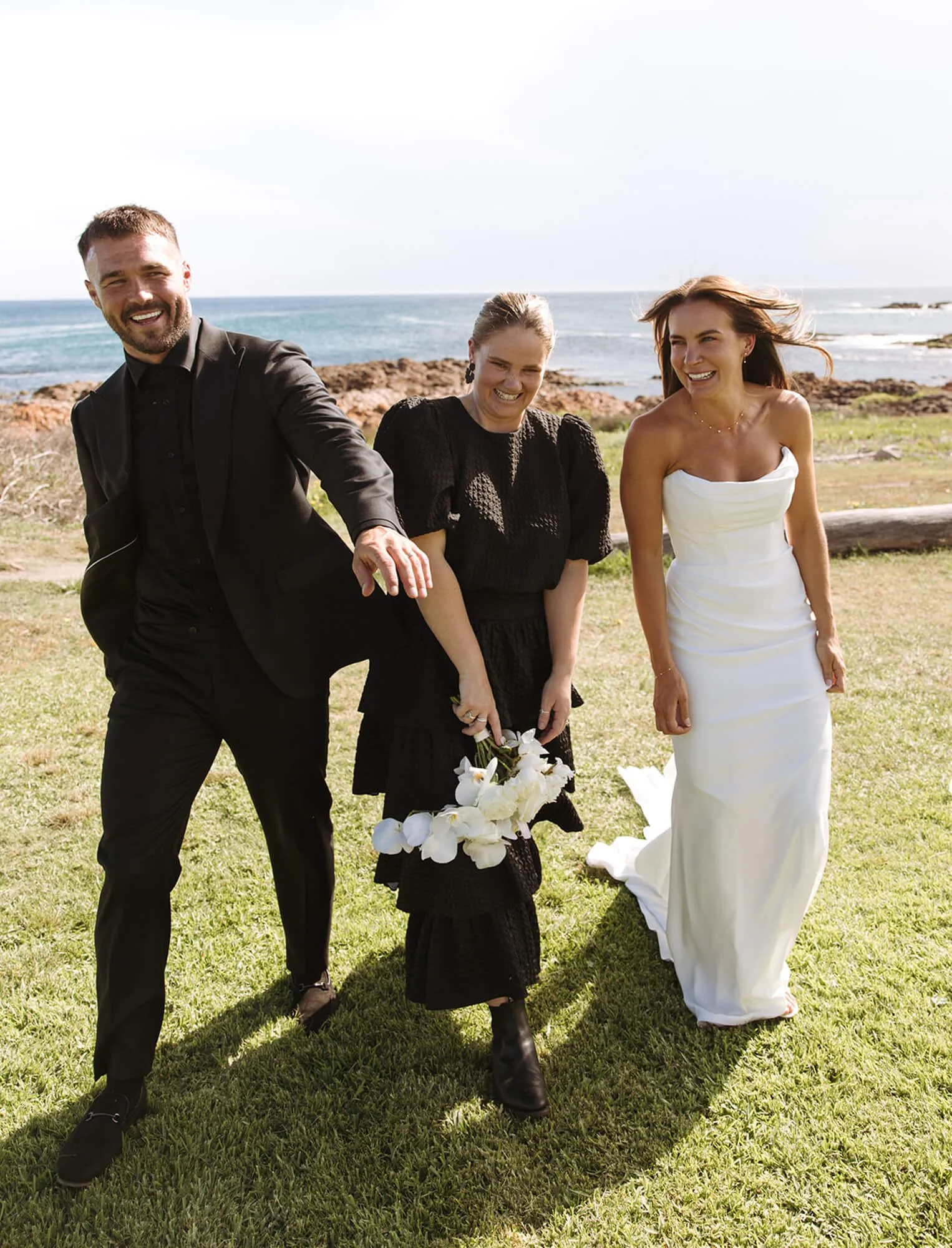 Three people at a beachside event, with the ocean in the background, smiling and walking on grass. One man in a black suit, a woman in a black dress holding a white flower bouquet, and another woman in a white strapless dress.