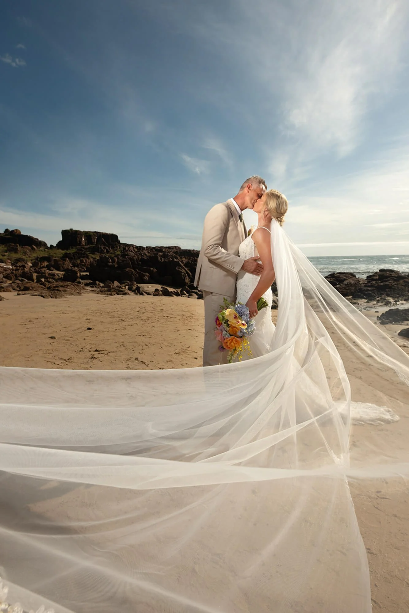 A bride and groom sharing a kiss on a beach, with the bride holding a bouquet of flowers and their wedding gown's veil flowing in the breeze.
