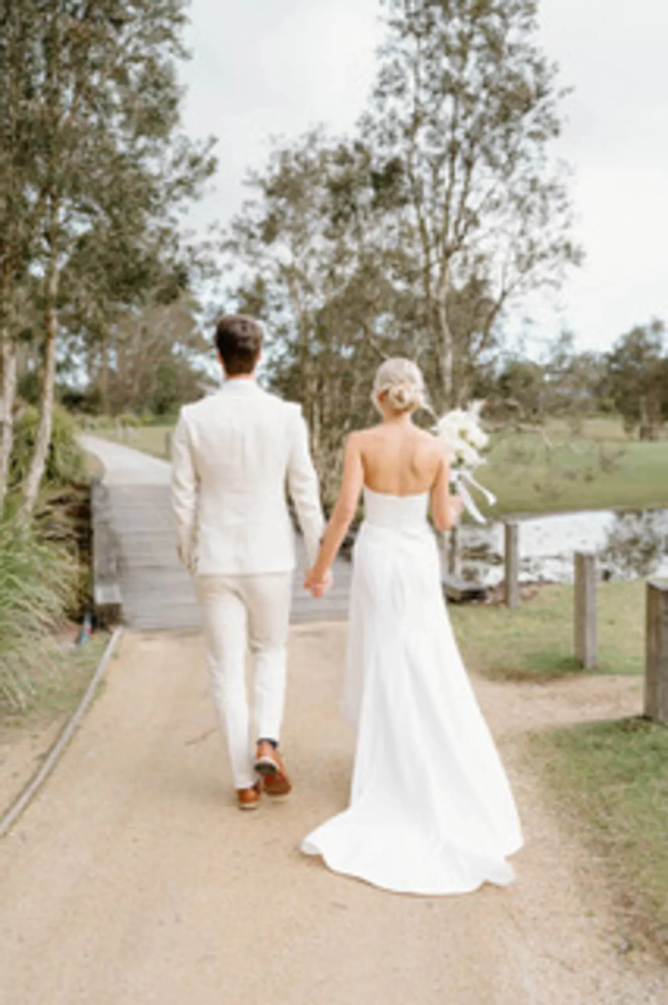 A bride and groom walking hand in hand along a path near a pond, with trees and greenery in the background, on their wedding day.