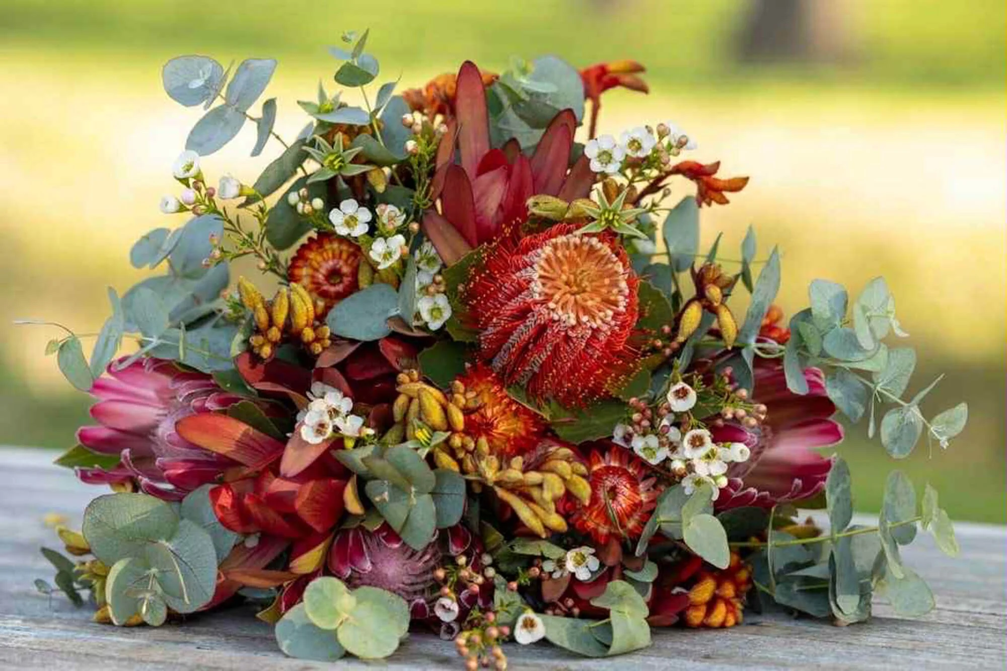 Colorful bouquet of red, orange, pink, and white flowers with green foliage, placed on a wooden surface with a blurred outdoor background.