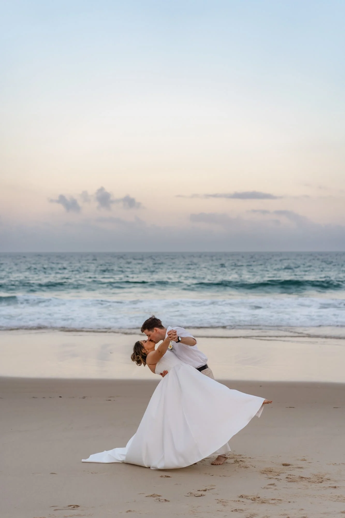 A couple dancing on the beach during sunset, with the ocean and sky in the background.