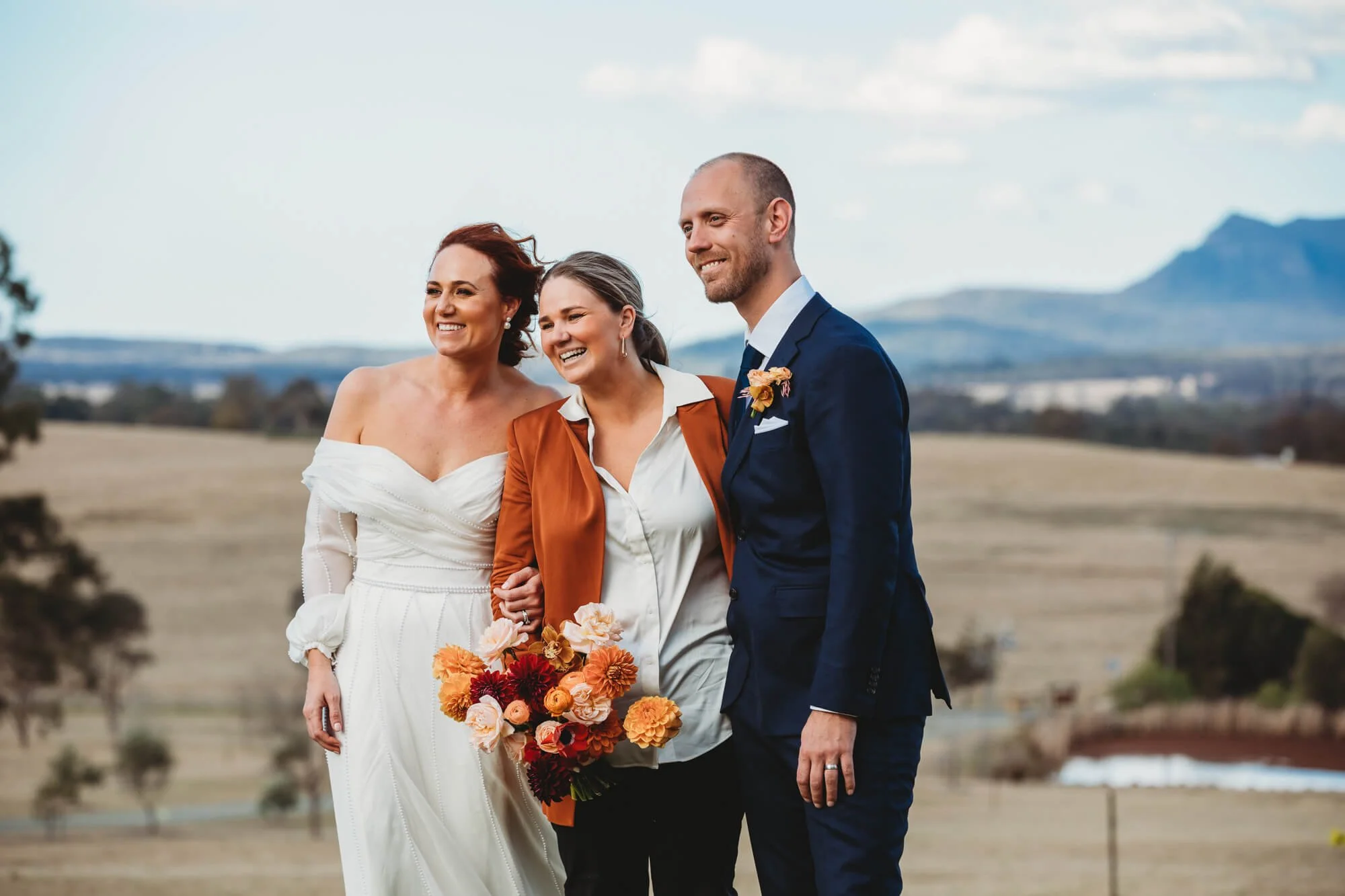 Three people at a wedding outdoors, two women and one man, smiling and holding a bouquet of flowers, with rolling hills and mountains in the background.
