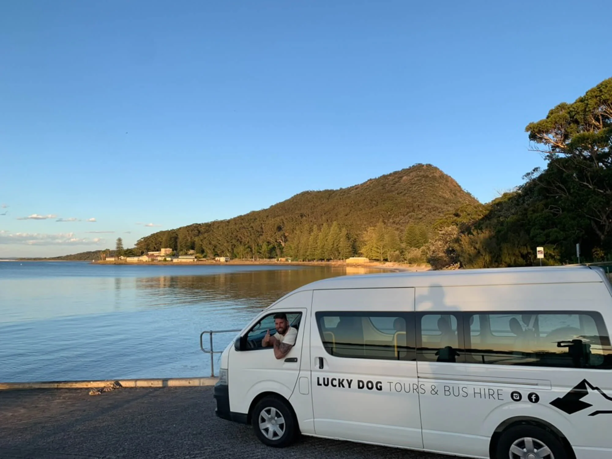 A man sitting in a white tour bus labeled "Lucky Dog Tours & Bus Hire" parked near a body of water with a hilly, forested landscape in the background during sunset.