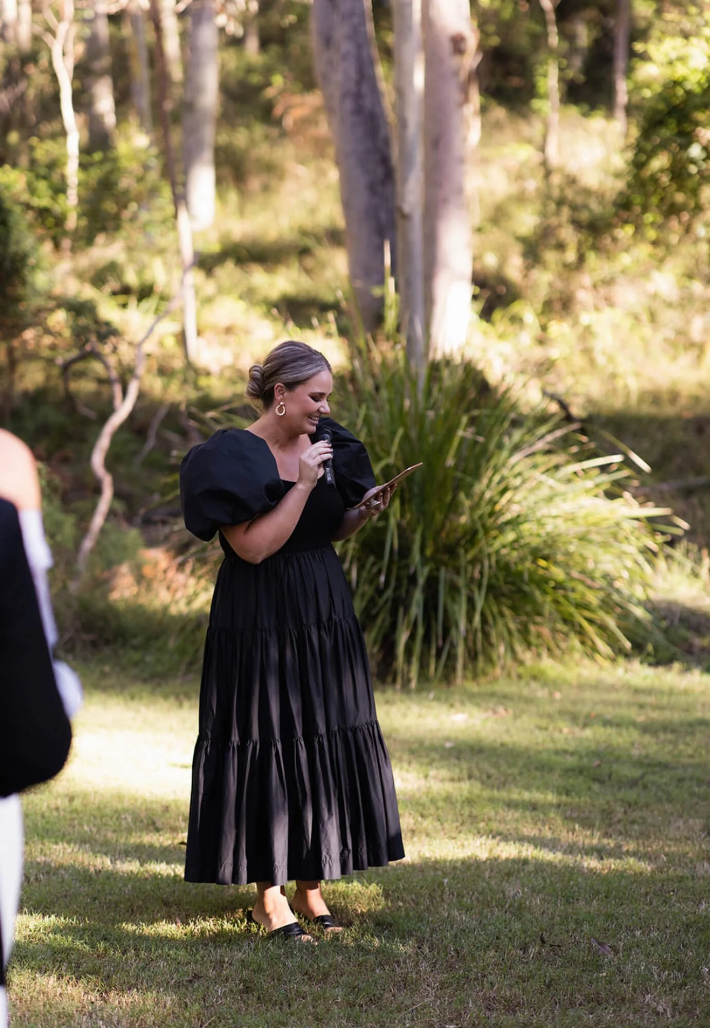 Woman in black dress holding microphone and reading from paper outdoors in a garden setting.