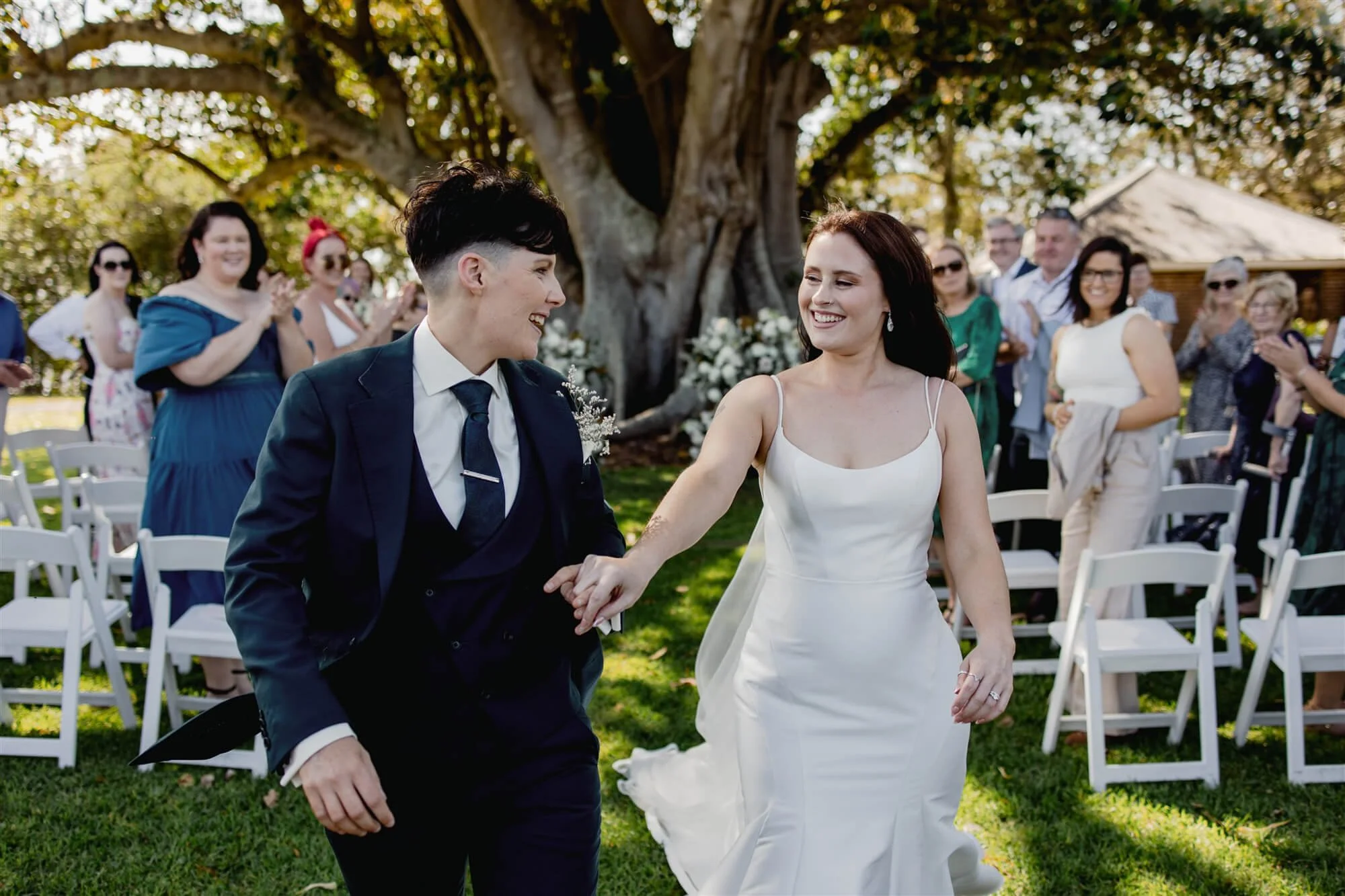 A bride and groom holding hands and smiling at each other at an outdoor wedding ceremony, with guests clapping in the background under a large tree.