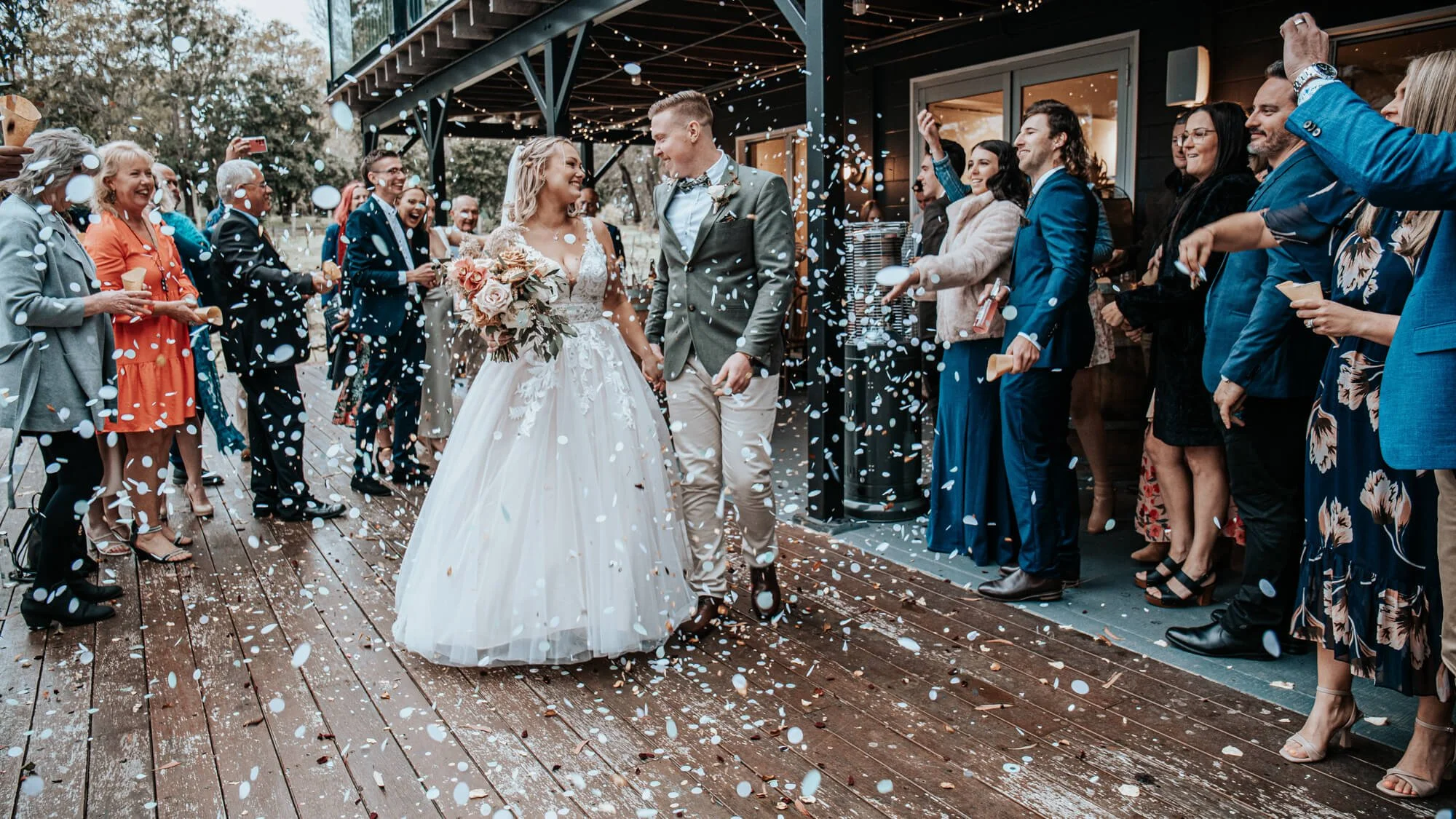 Bride and groom are walking hand in hand through a confetti shower surrounded by guests at their wedding celebration.