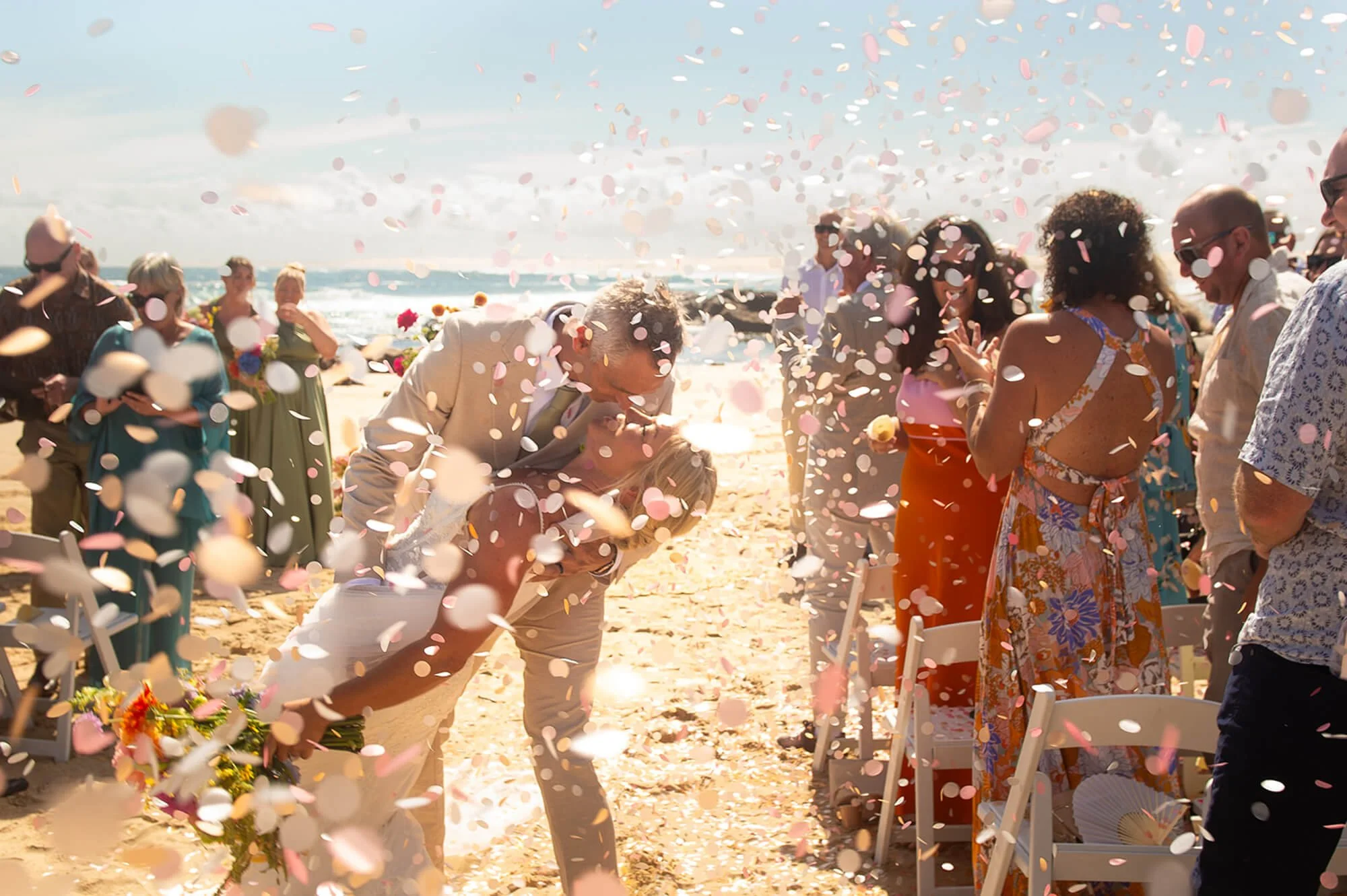 Couple kisses at a beach wedding with guests celebrating as pink and white confetti falls.