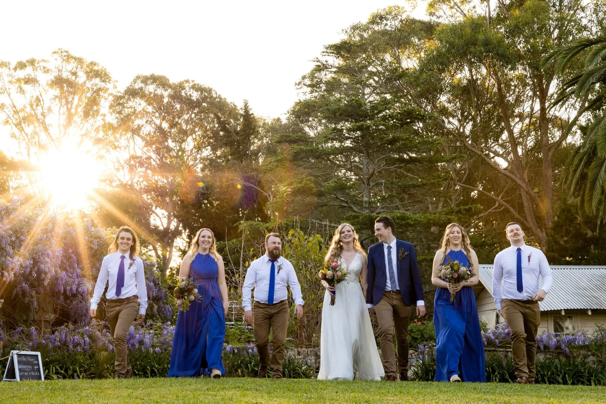 A group of seven people at a wedding, including the bride in a white dress holding a bouquet, and six other people in formal attire, walking on a lawn during sunset with trees and a building in the background.