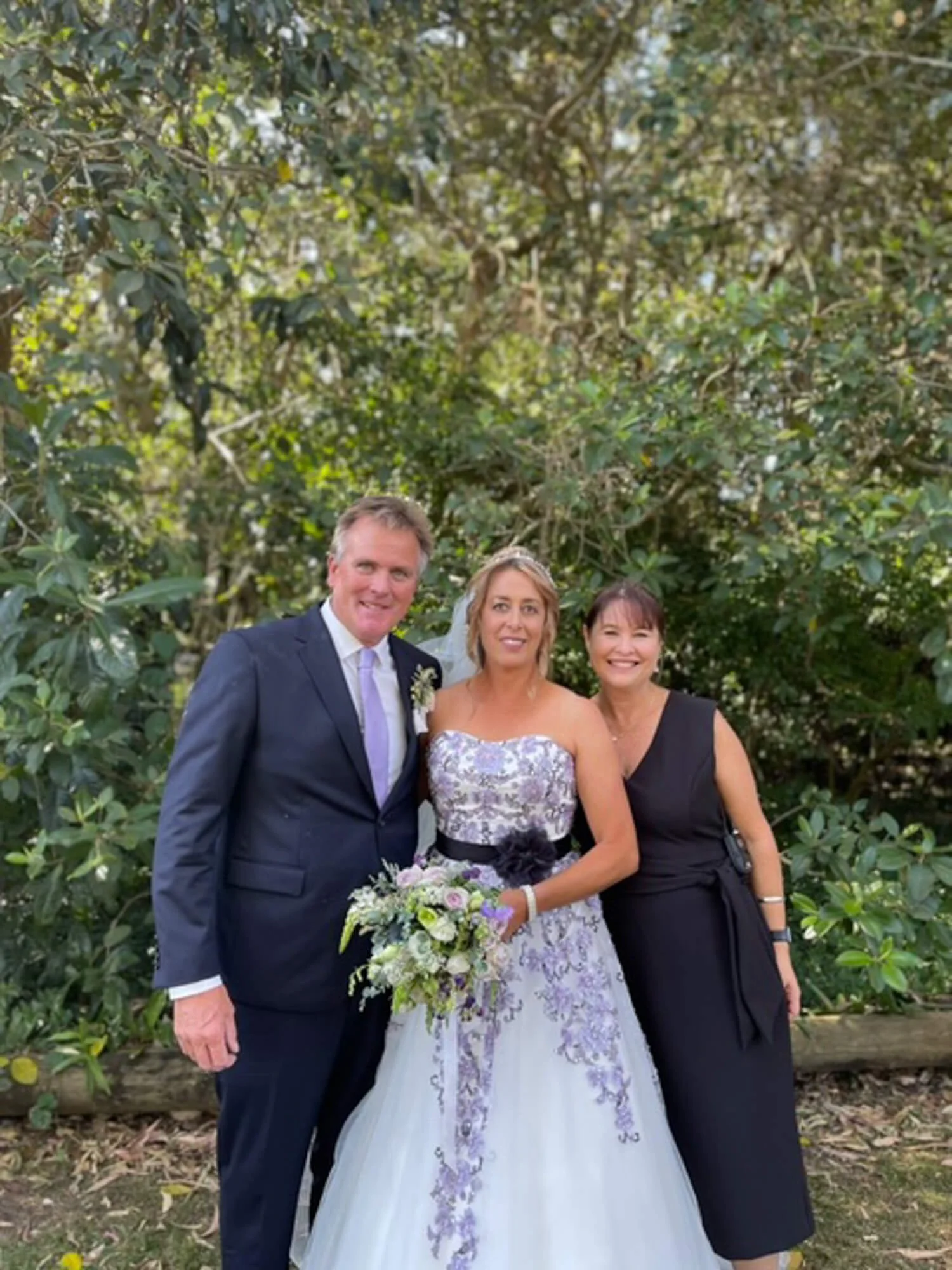 A bride in a white gown holding a bouquet, a groom in a suit, and a woman in a black dress standing in front of green foliage at a wedding.