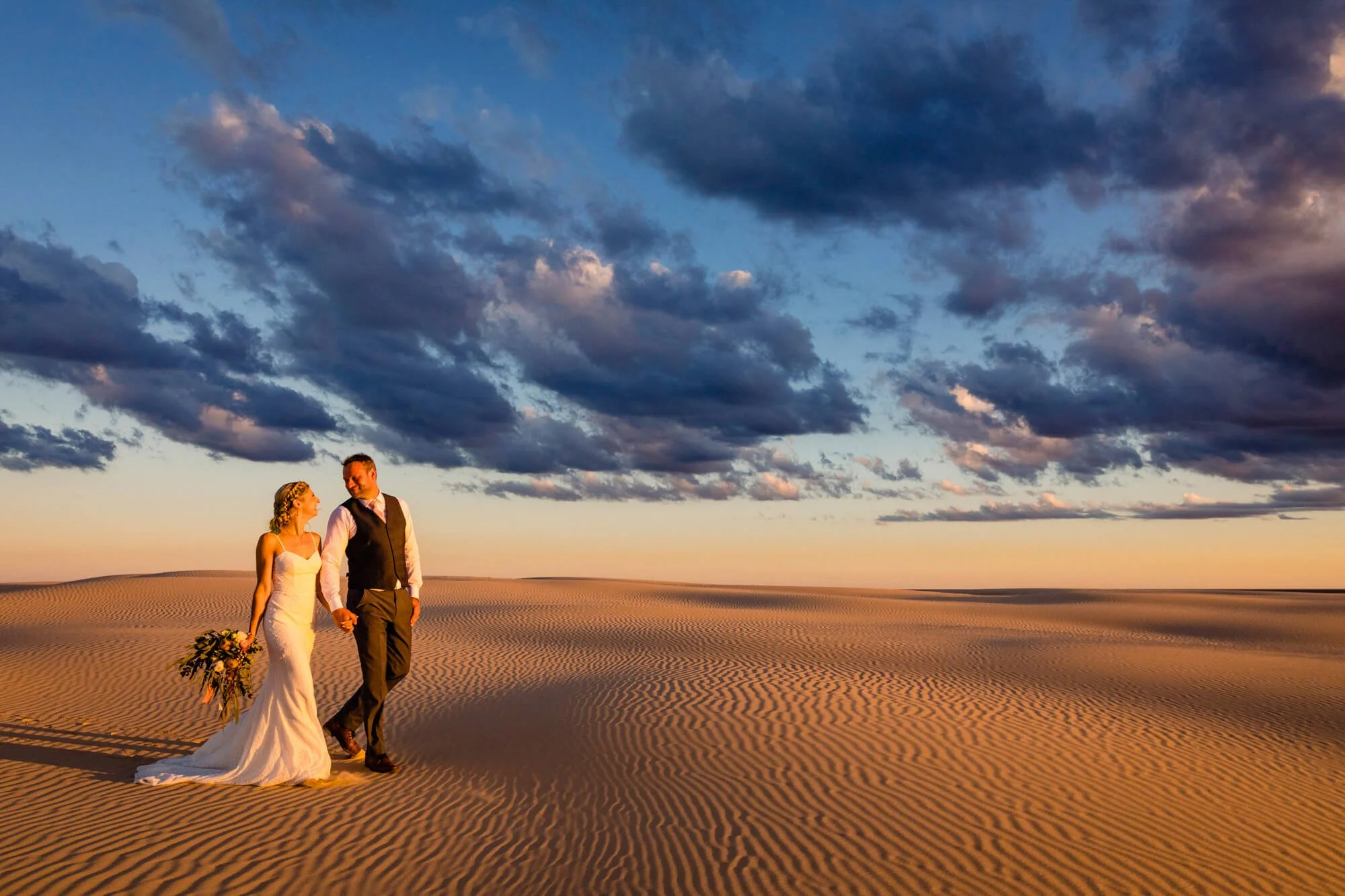 A bride and groom walking hand in hand on a desert dune at sunset, with dramatic clouds in the sky.