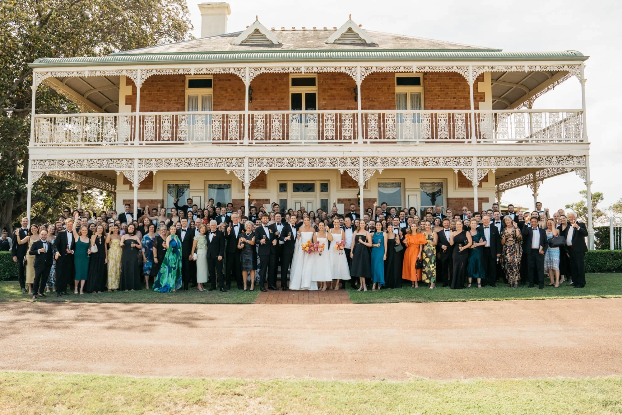 A large group of wedding guests and the bride and groom, standing outside in front of a historic two-story building with ornate white lace trim balcony, celebrating the wedding.