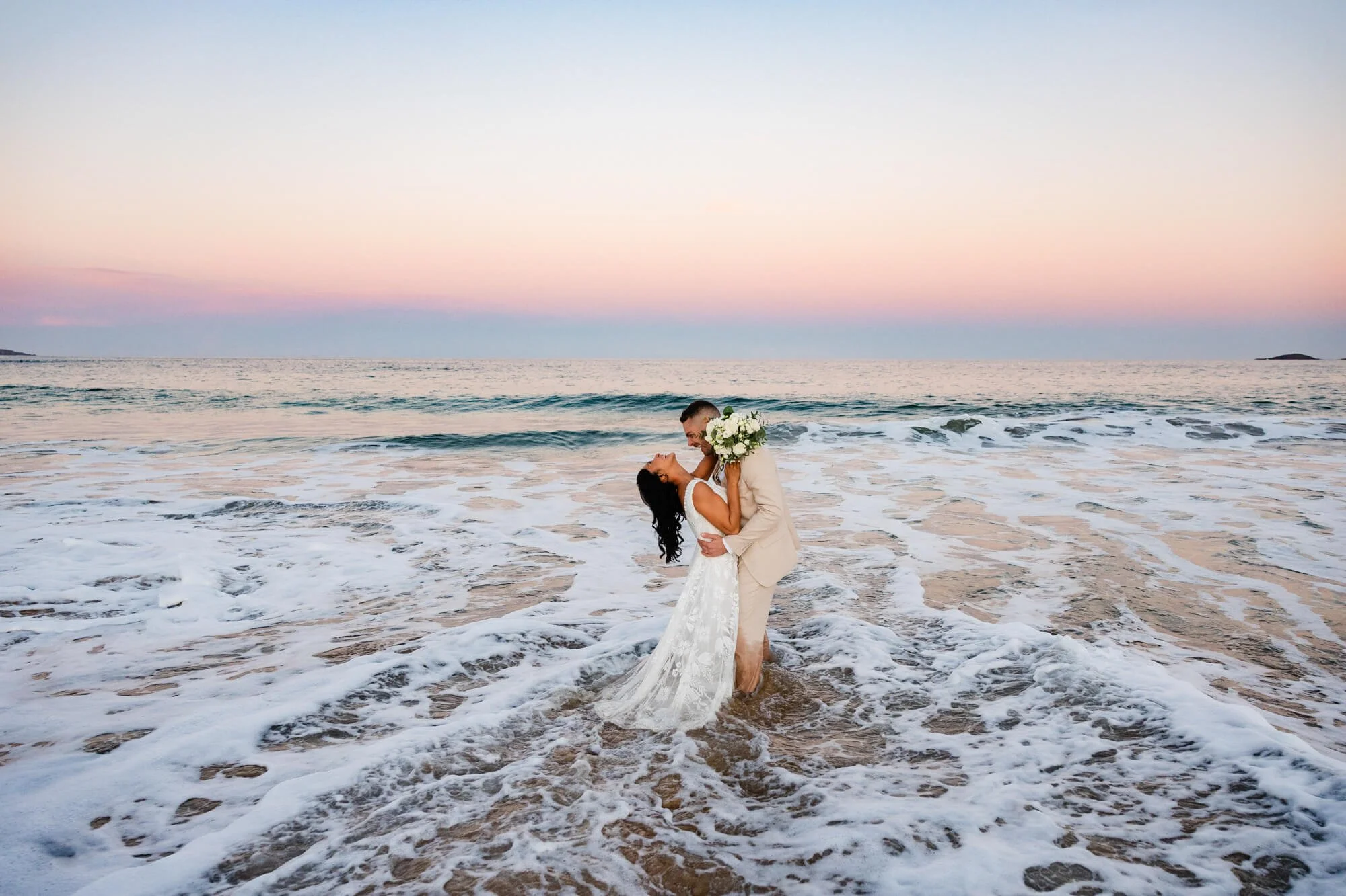 A couple dressed in wedding attire standing in the ocean at sunset, sharing a kiss, with the bride holding a bouquet of white flowers.