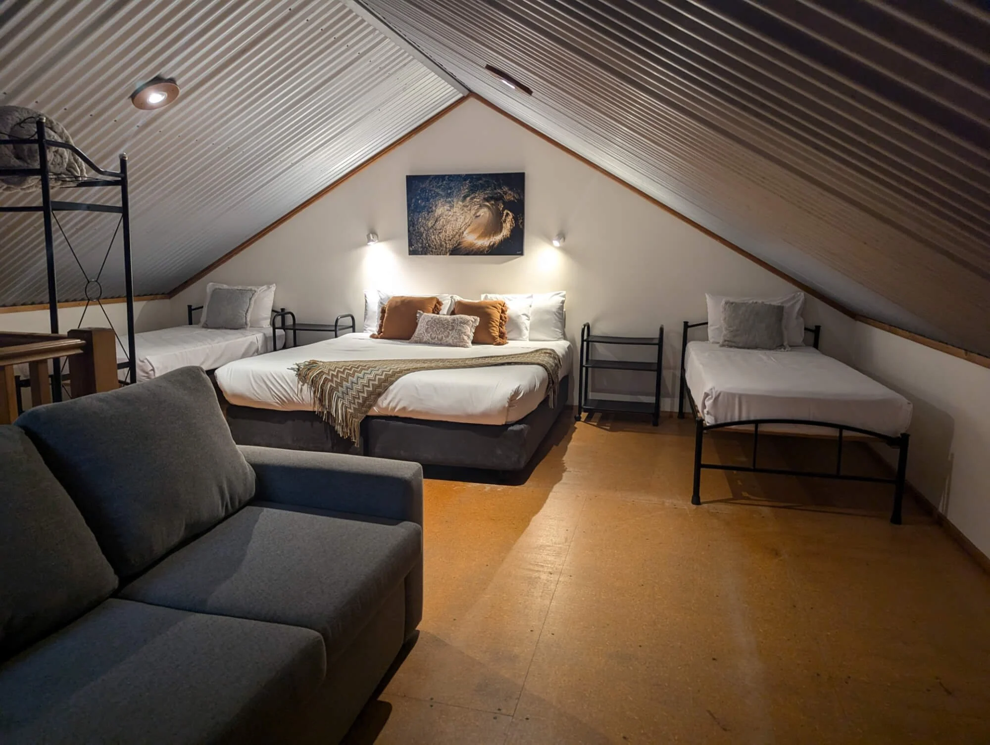 Attic bedroom with three beds, a gray sofa, and wall art of a wave, with warm lighting and wooden flooring.