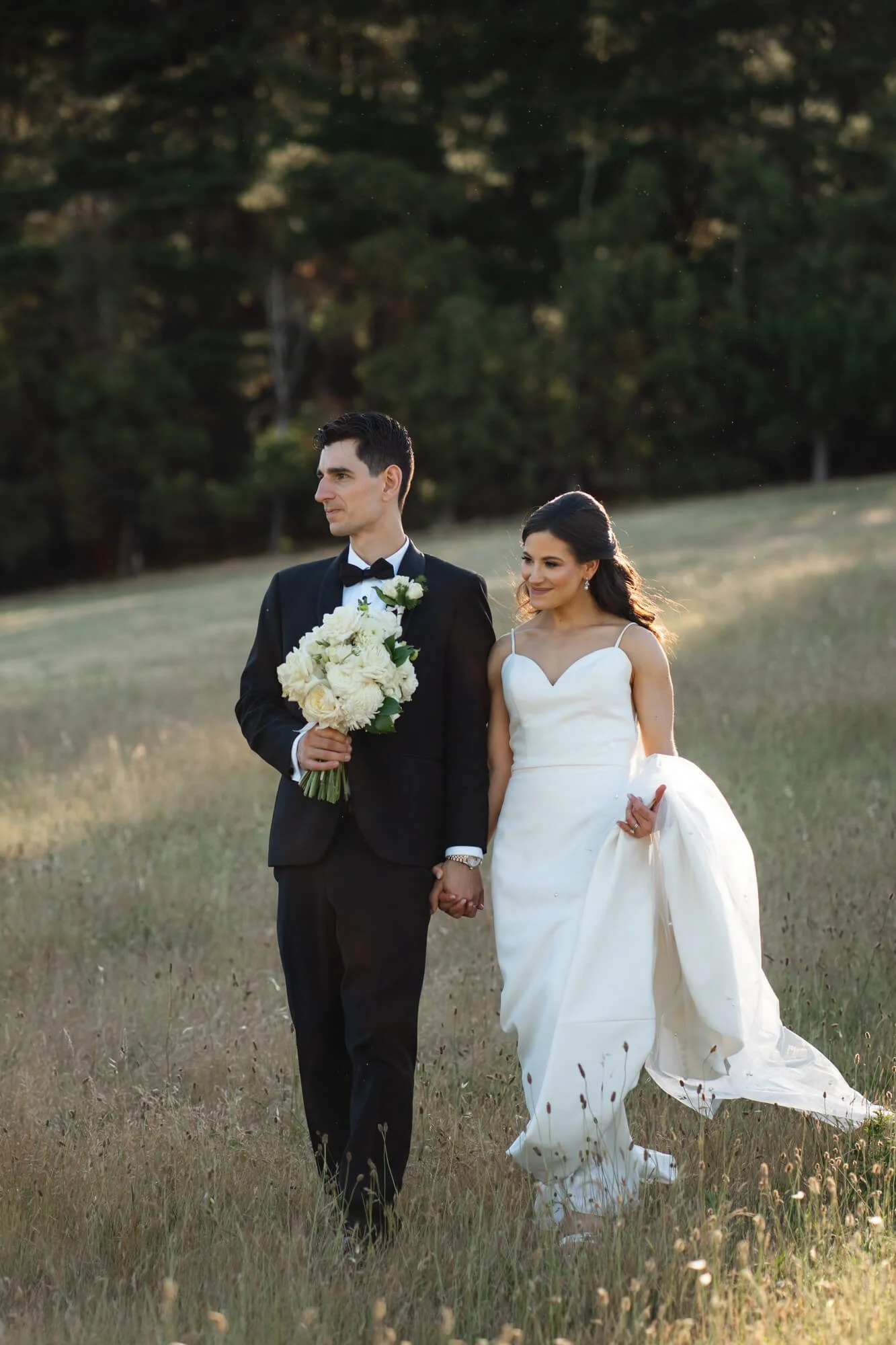 Bride and groom walking hand in hand in a grassy field during sunset, the groom in a black tuxedo holding a bouquet of white flowers, the bride in a white wedding gown carrying her dress, both smiling.