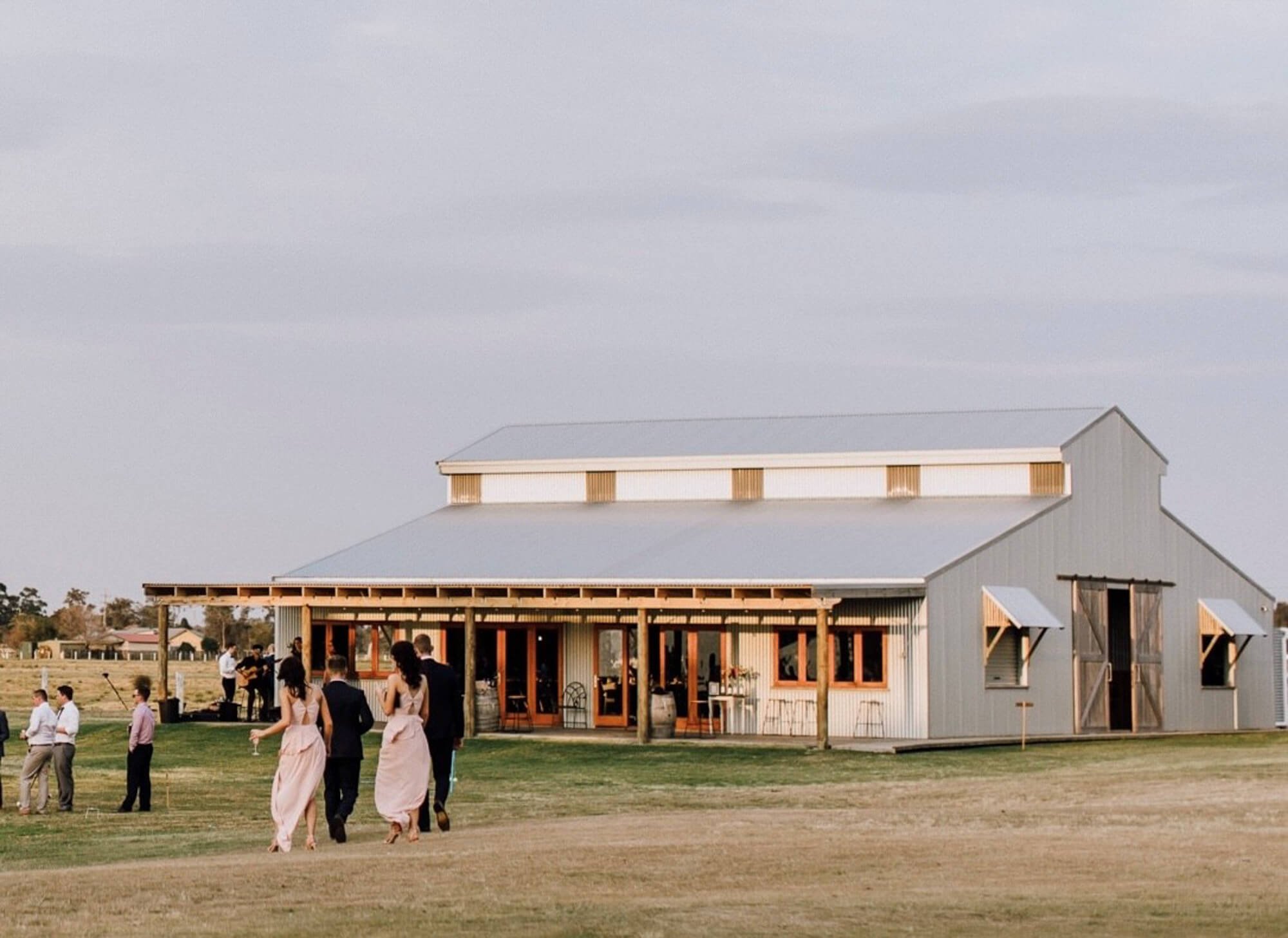 A group of people dressed in formal attire walking towards a large, rustic barn with a covered porch, set in a grassy field during the daytime.