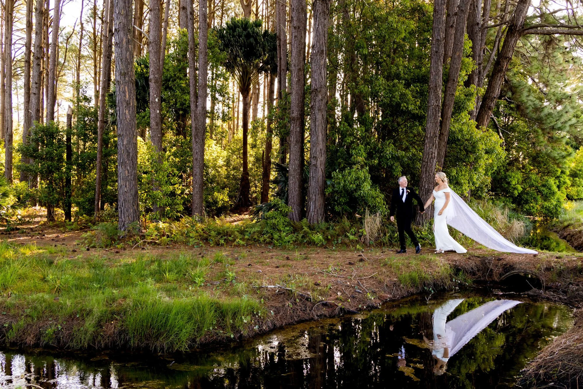 A bride and groom walking hand in hand in a forested area near a small creek, with tall trees and sunlight filtering through the leaves.