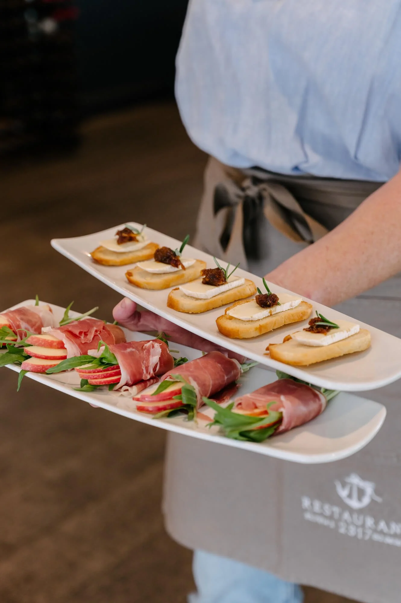 A person holding two rectangular white plates with appetizers. The top plate has toasted baguette slices topped with cheese and a small meat garnish. The bottom plate has rolled slices of prosciutto with greens and sliced apples.