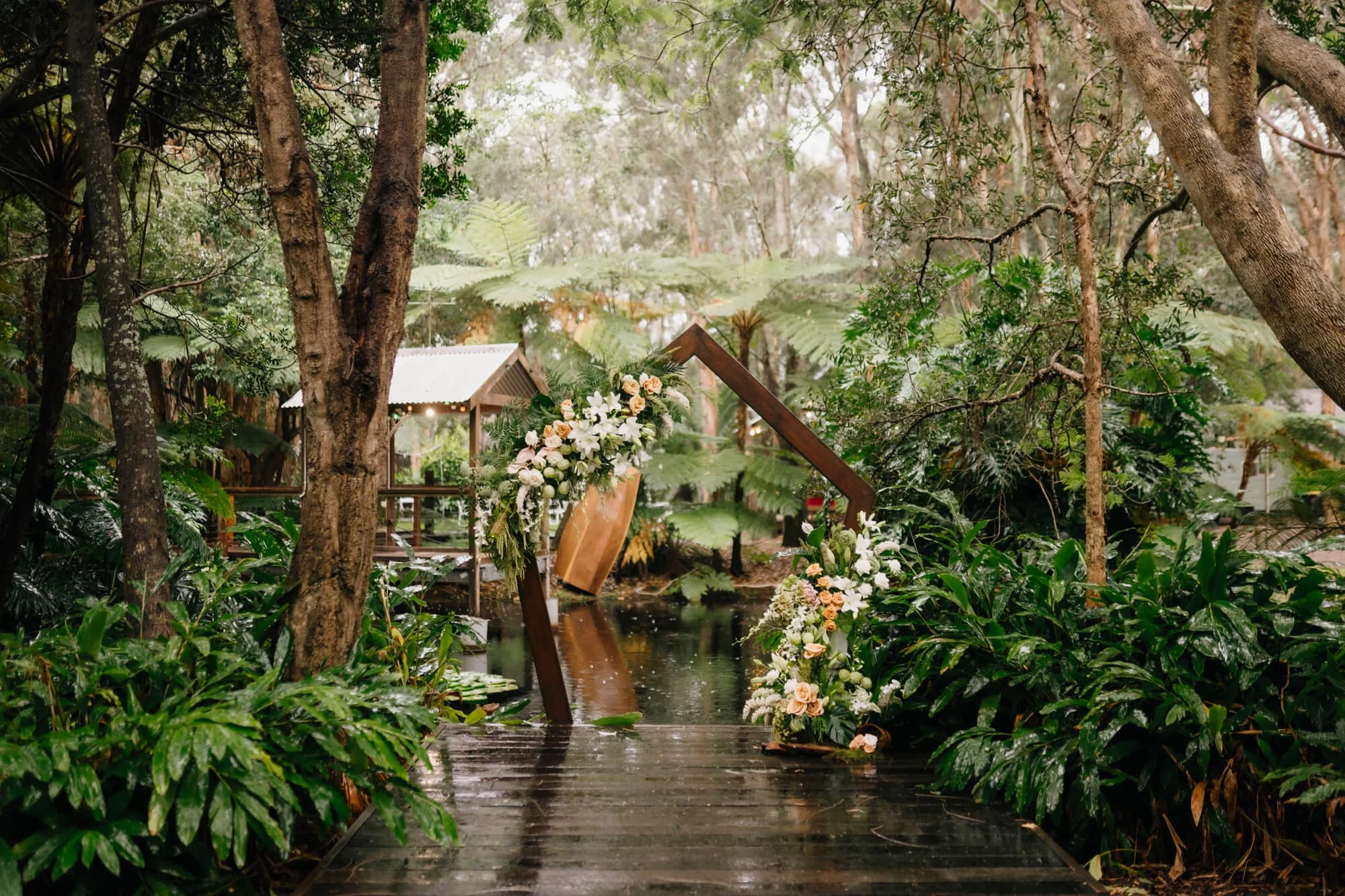 Outdoor wedding ceremony setting in a lush, green forest with a floral arch over a wooden pathway.