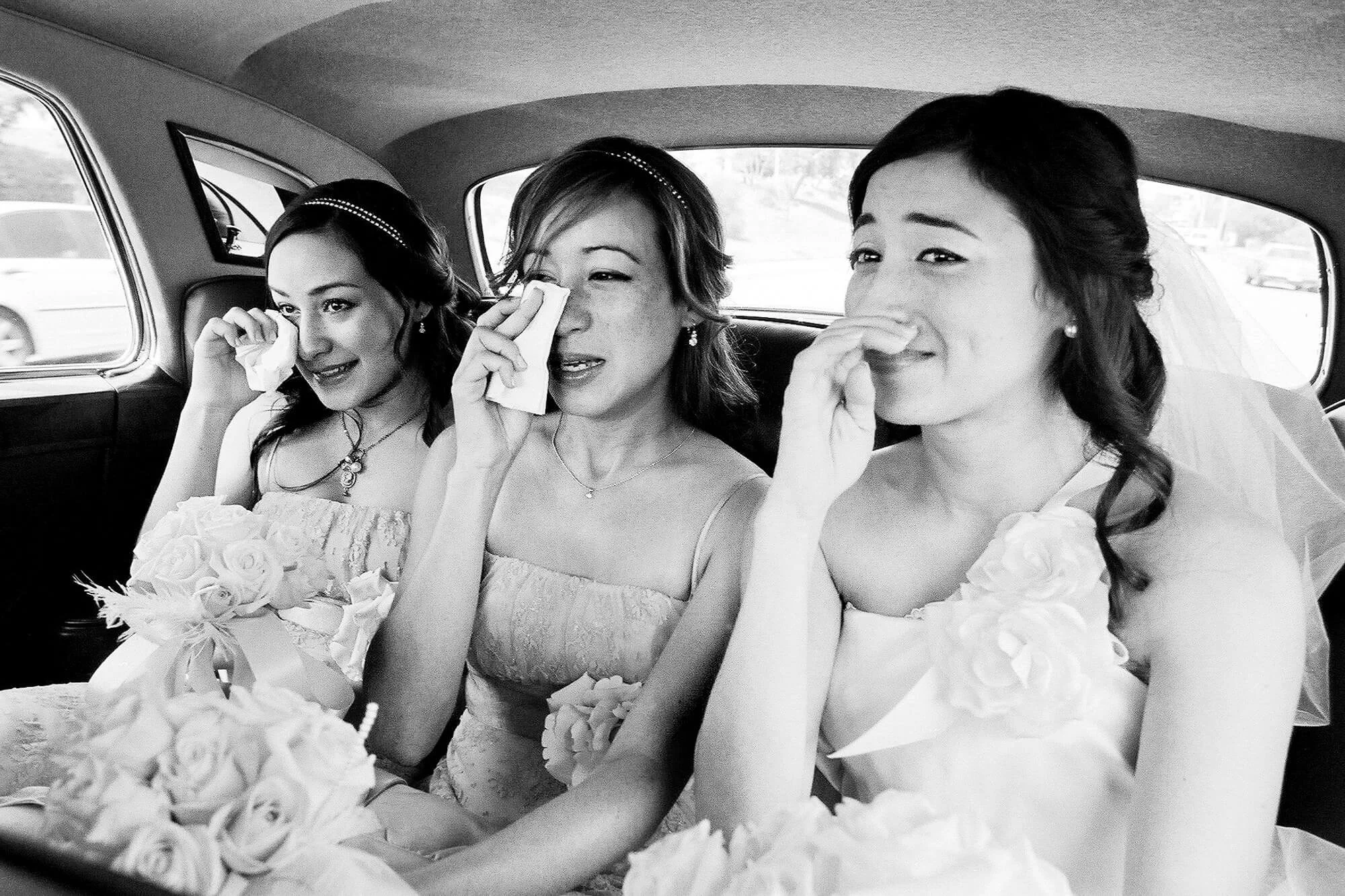 Three women sitting in the back of a car, appearing emotional and wiping tears, dressed in wedding attire.