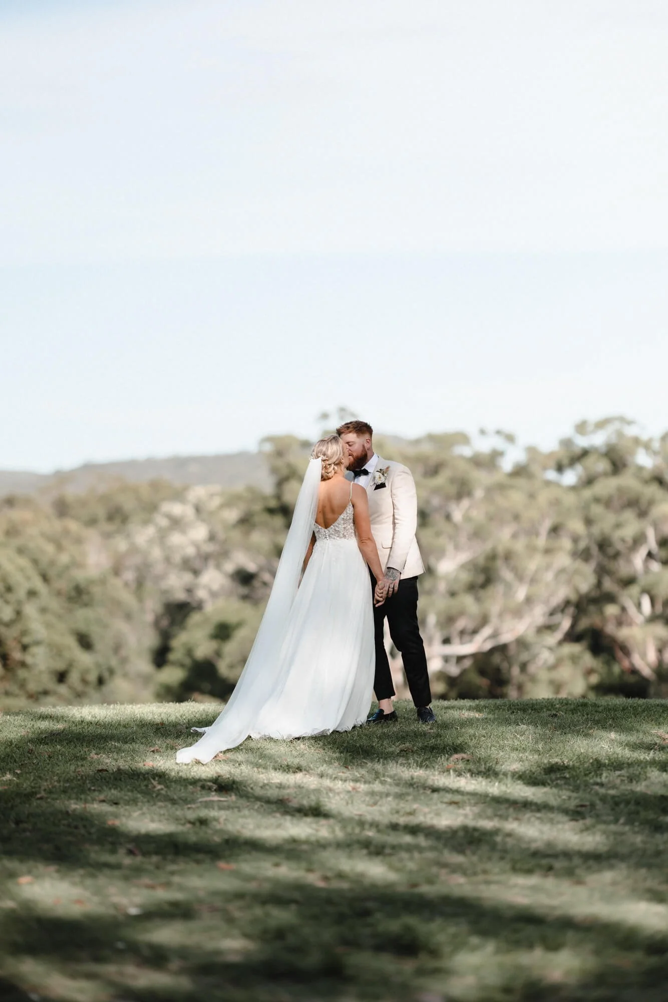 A bride and groom kiss outdoors on a grassy hill with a blurred treeline in the background.