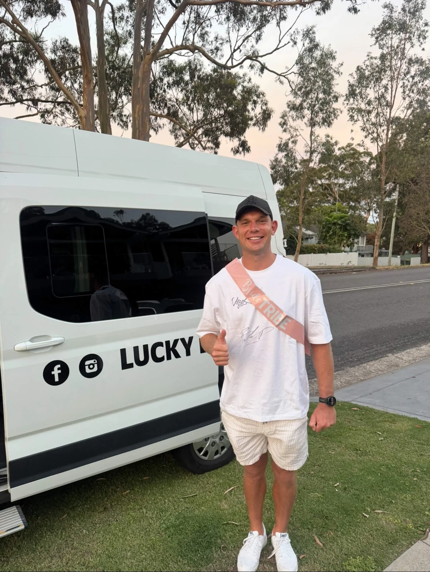 A smiling man in a white t-shirt, beige shorts, white sneakers, and a black cap standing outdoors on the grass, giving a thumbs-up gesture beside a white van with social media icons and the word "LUCKY" on it. The man is wearing a pink sash that says