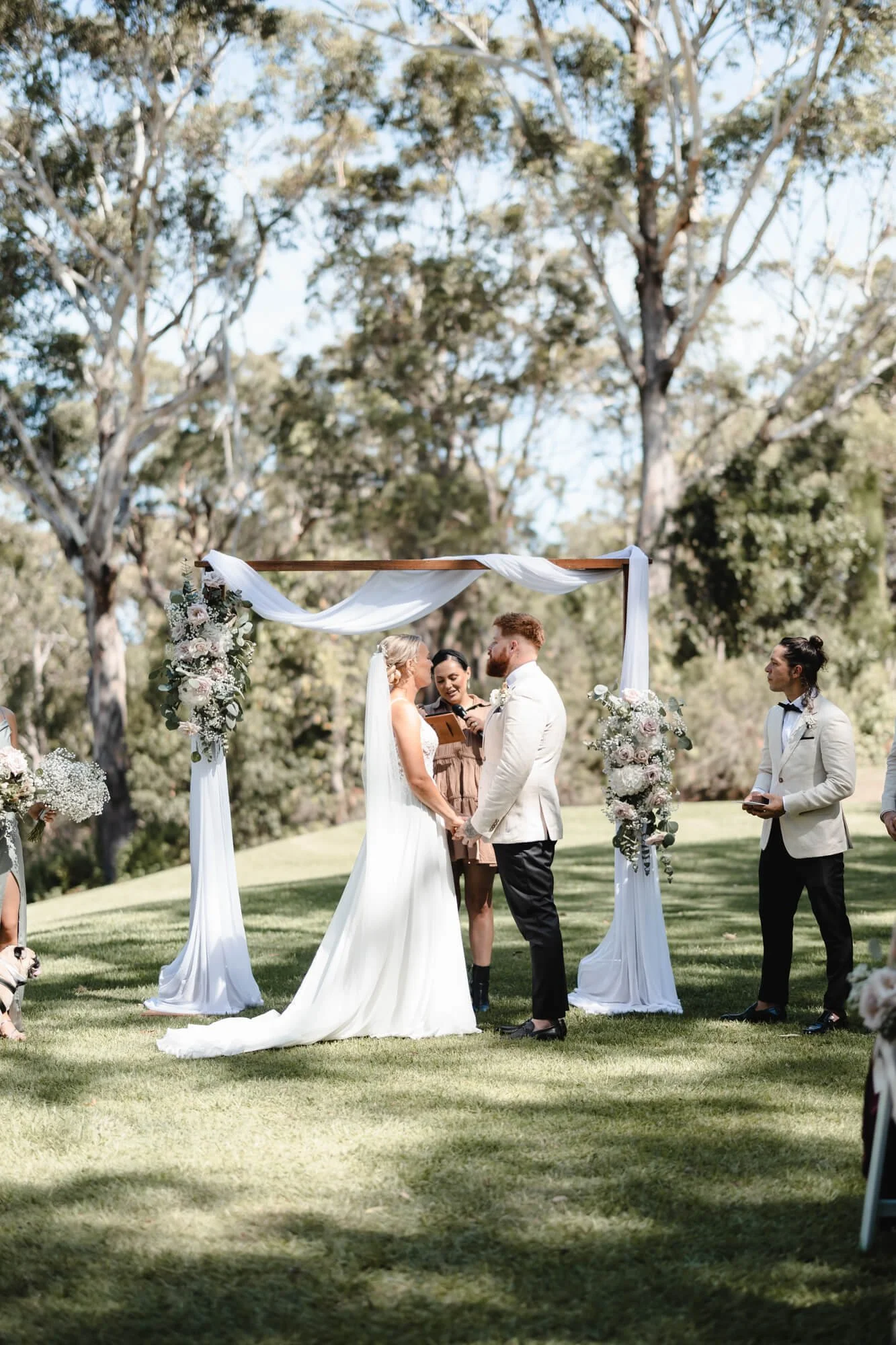A couple getting married outdoors under a decorated wedding arch, with a officiant and officiant's assistant standing nearby, surrounded by trees and greenery.