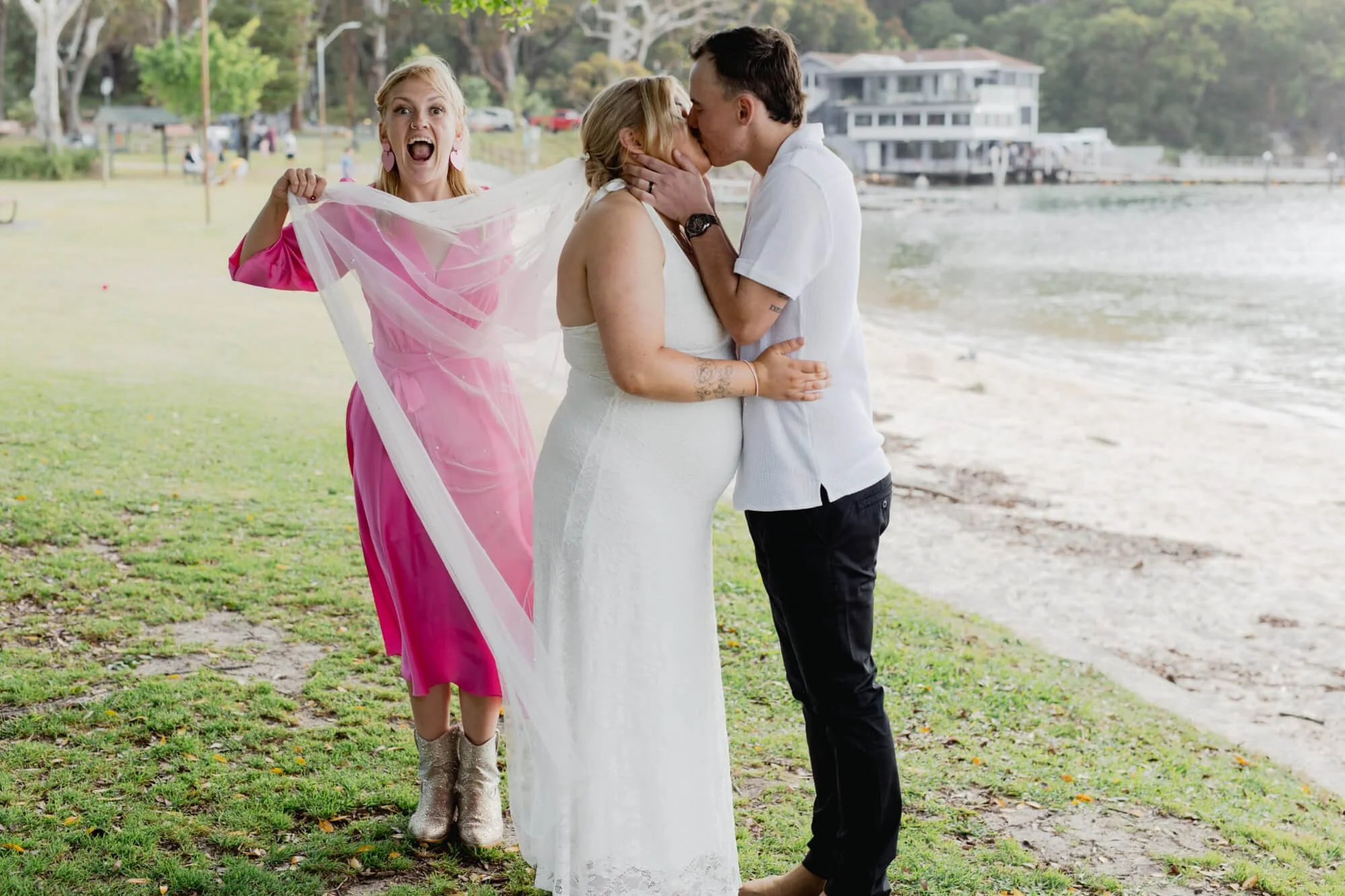 A couple is kissing by the beach while a woman in a pink dress and glittery boots holds a veil and reacts happily.