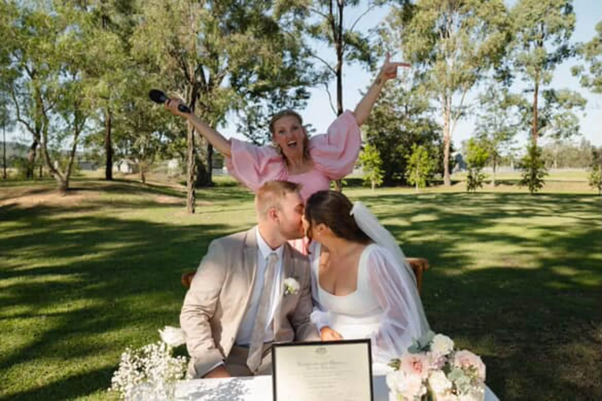 A wedding couple kissing outdoors at a park with trees in the background, sitting at a table decorated with flowers and a framed document, with a woman in a pink dress celebrating enthusiastically behind them.